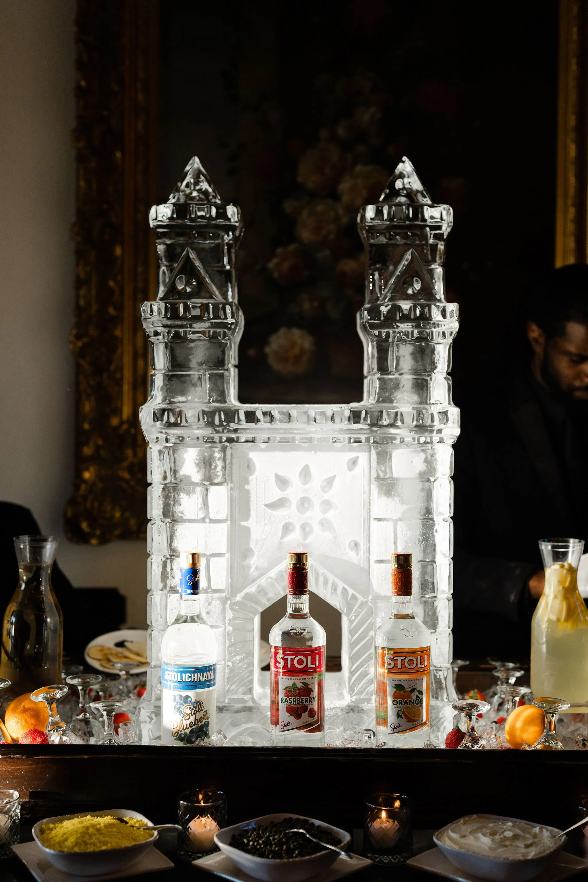 Ice sculpture of a castle with bottles of Stoli vodka in front, surrounded by fruits and food from a buffet table.