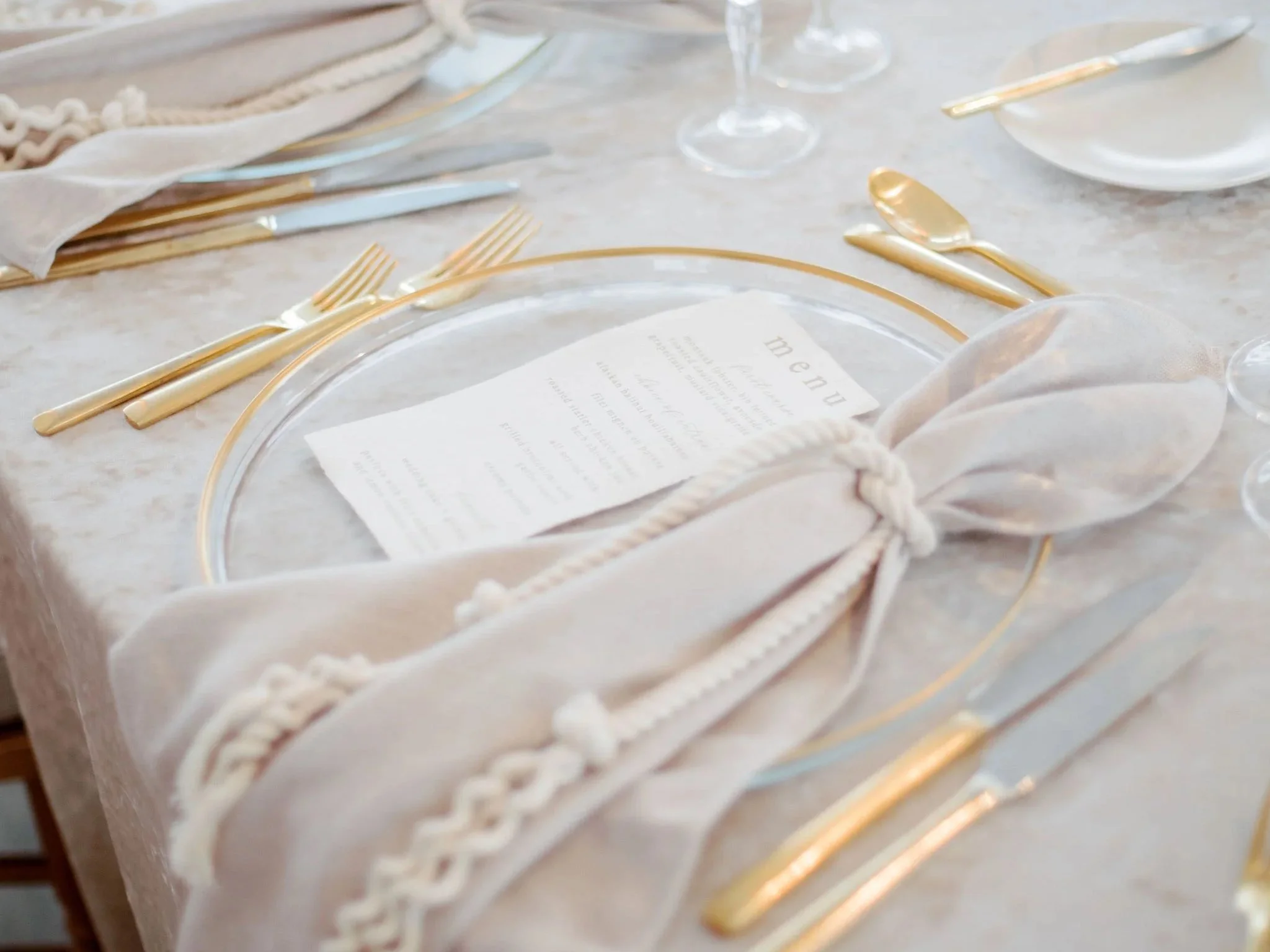 Neutral-toned linens, gold flatware, and handcrafted deckled edge menus come together in a refined coastal tablescape, accented with subtle nautical rope details.
