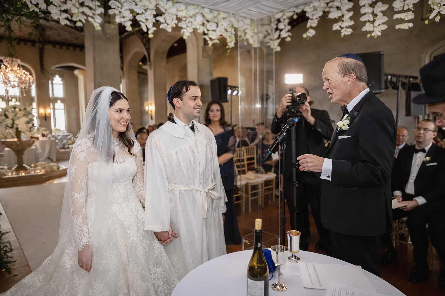 Jewish wedding ceremony with bride and groom in his kittel under a lucite and orchid chuppah during blessing at Hempstead House