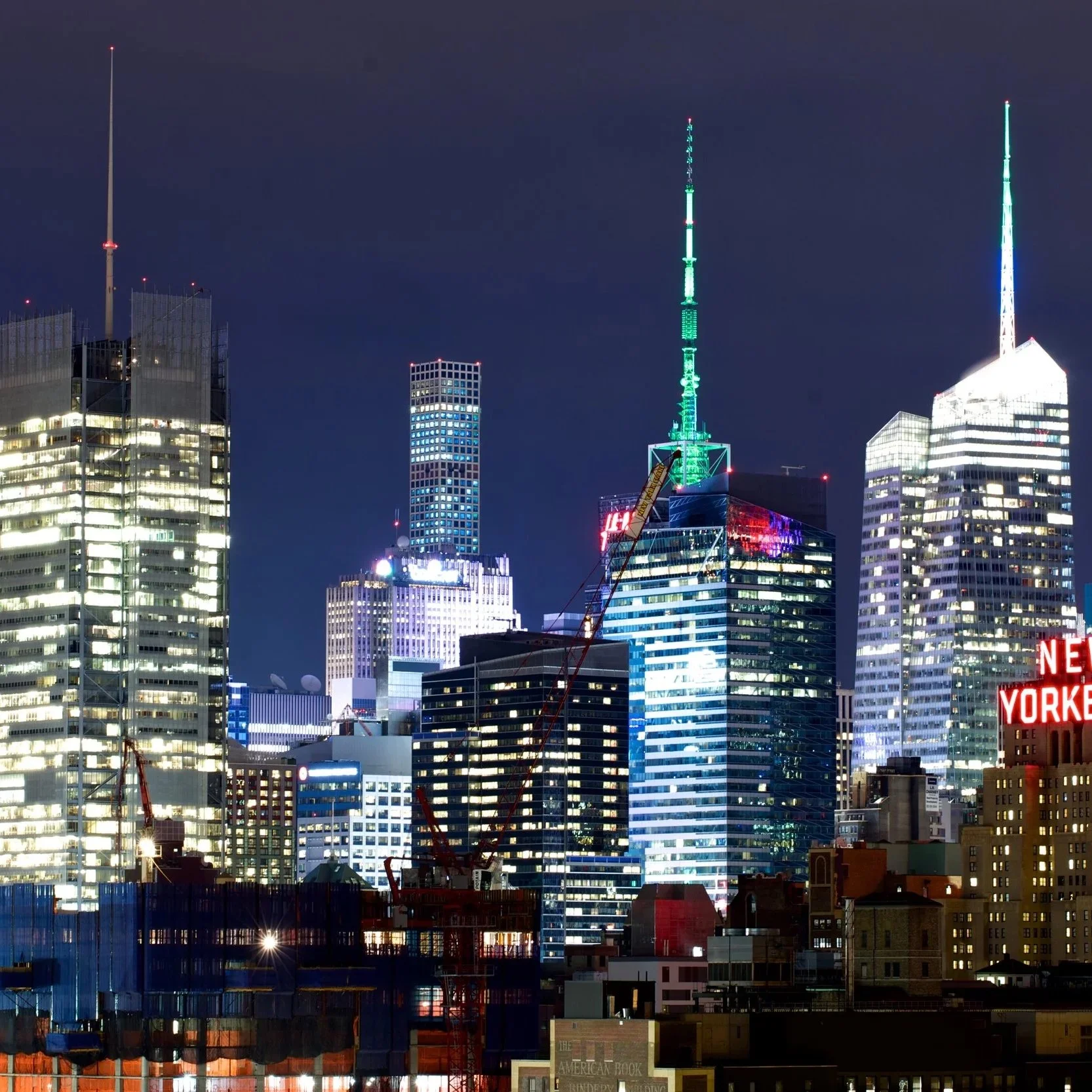 Nighttime view of the New York City skyline with illuminated skyscrapers, construction cranes, and the iconic One Vanderbilt building.