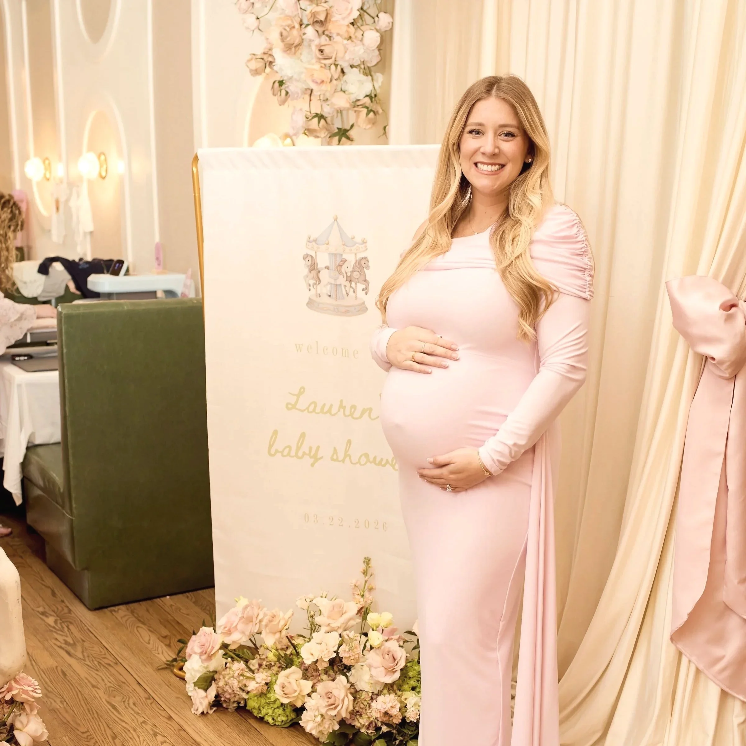 Lauren’s baby shower at Arlo in Northport, NY.  Behind her there is a decorated backdrop with flowers and a carousel theme. There are pink and white flowers arranged on the floor and in the background, and a sign with the words 'lauren' and 'baby sho