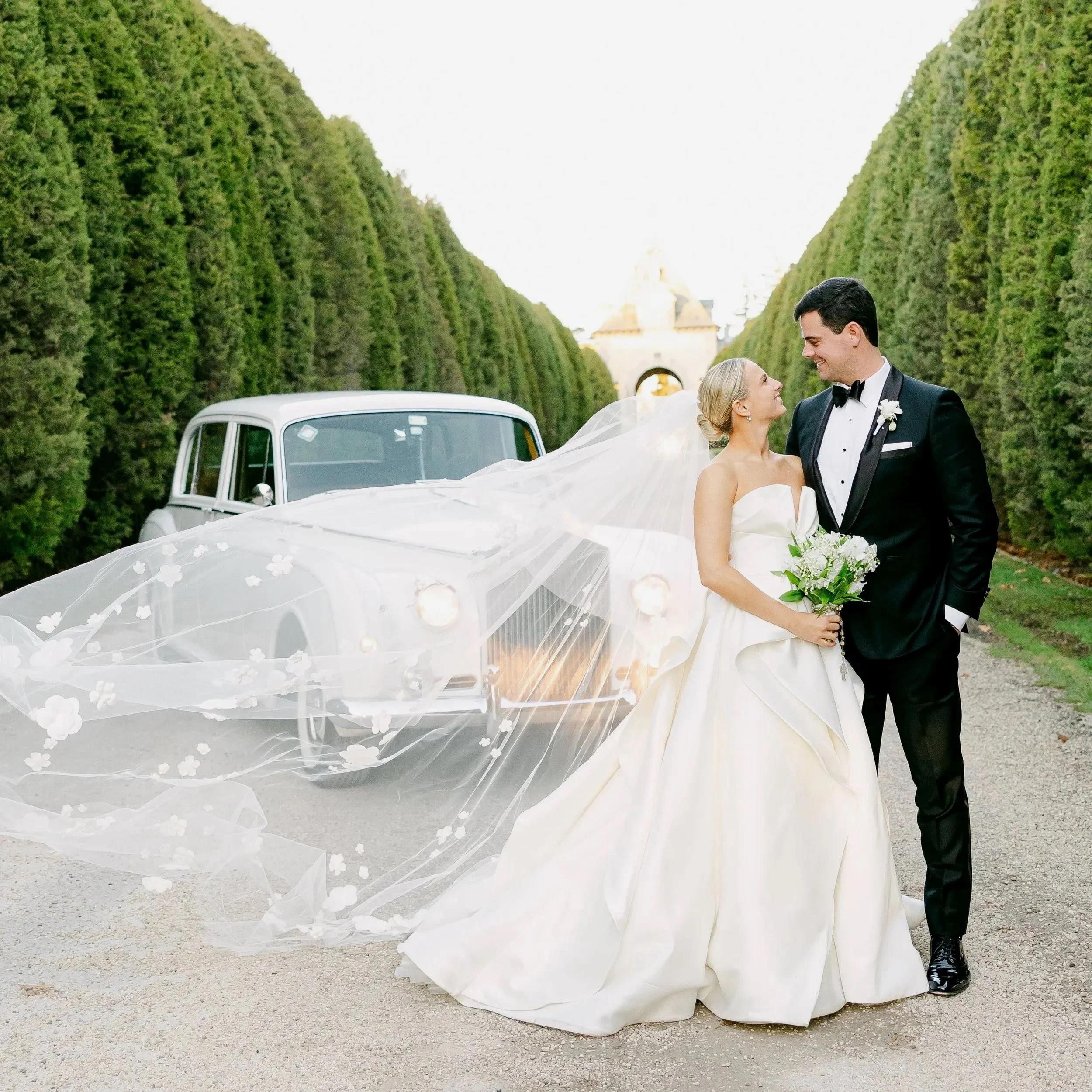 Bride with flowing veil poses with her groom in front of a vintage Rolls Royce in the driveway at Oheka Castle 