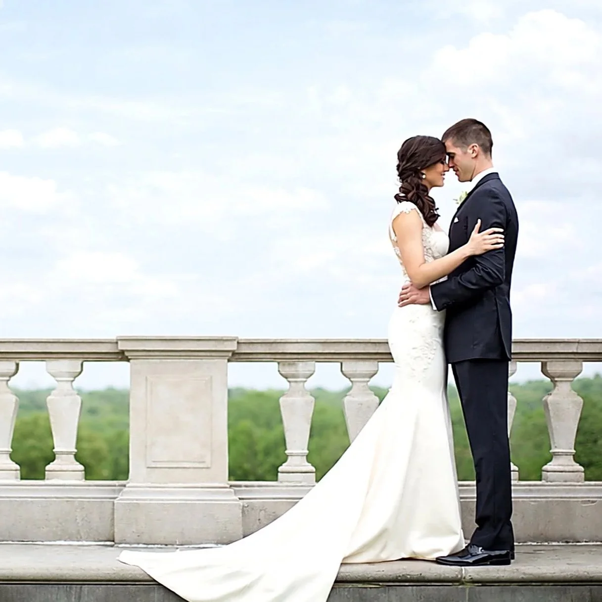 Bride and groom standing close together on a stone balcony, facing each other, with a scenic green landscape and sky in the background.