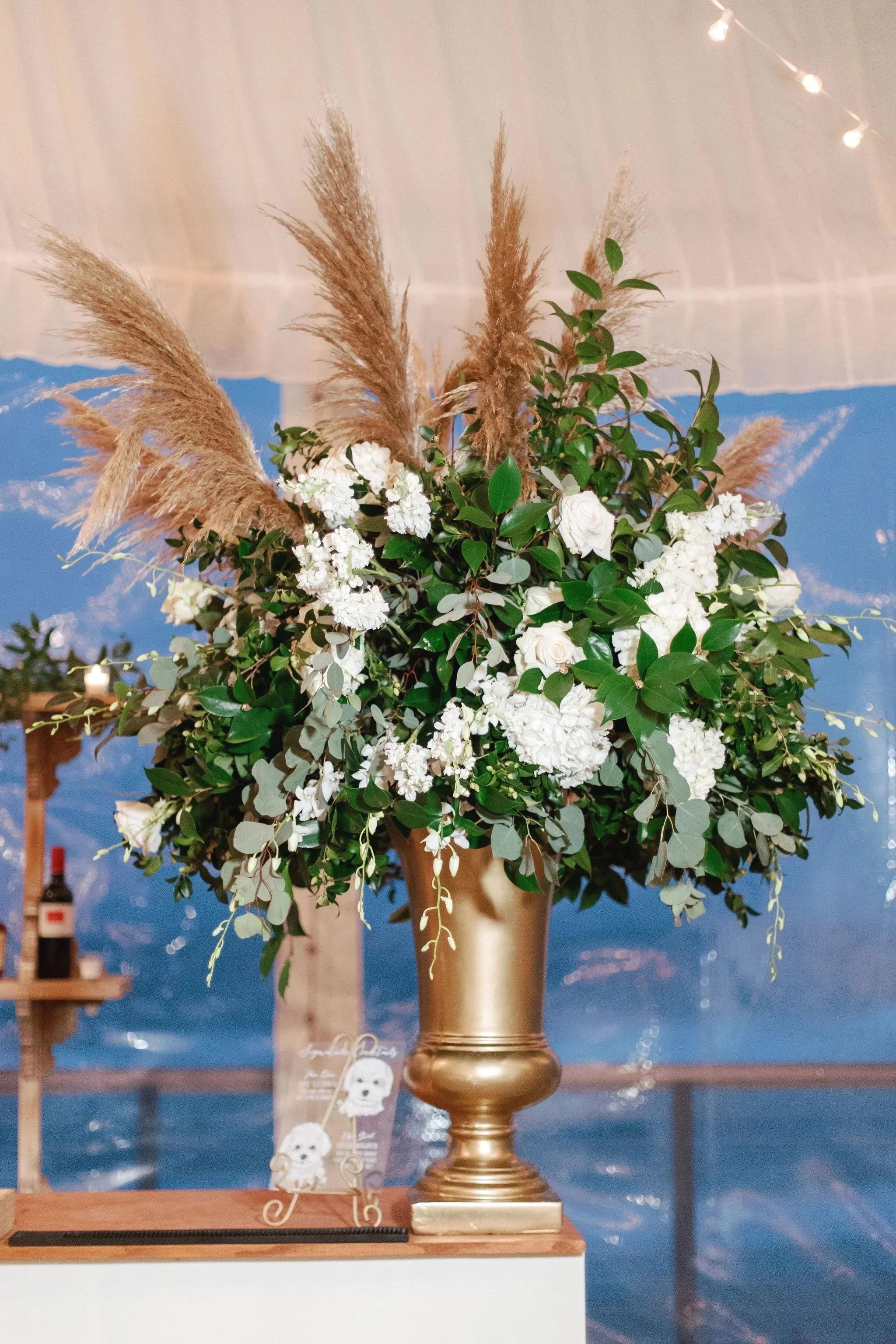 A floral arrangement in a gold vase with pampas grass, white roses, and greenery, placed on a table with a navy blue abstract backdrop and string lights overhead.