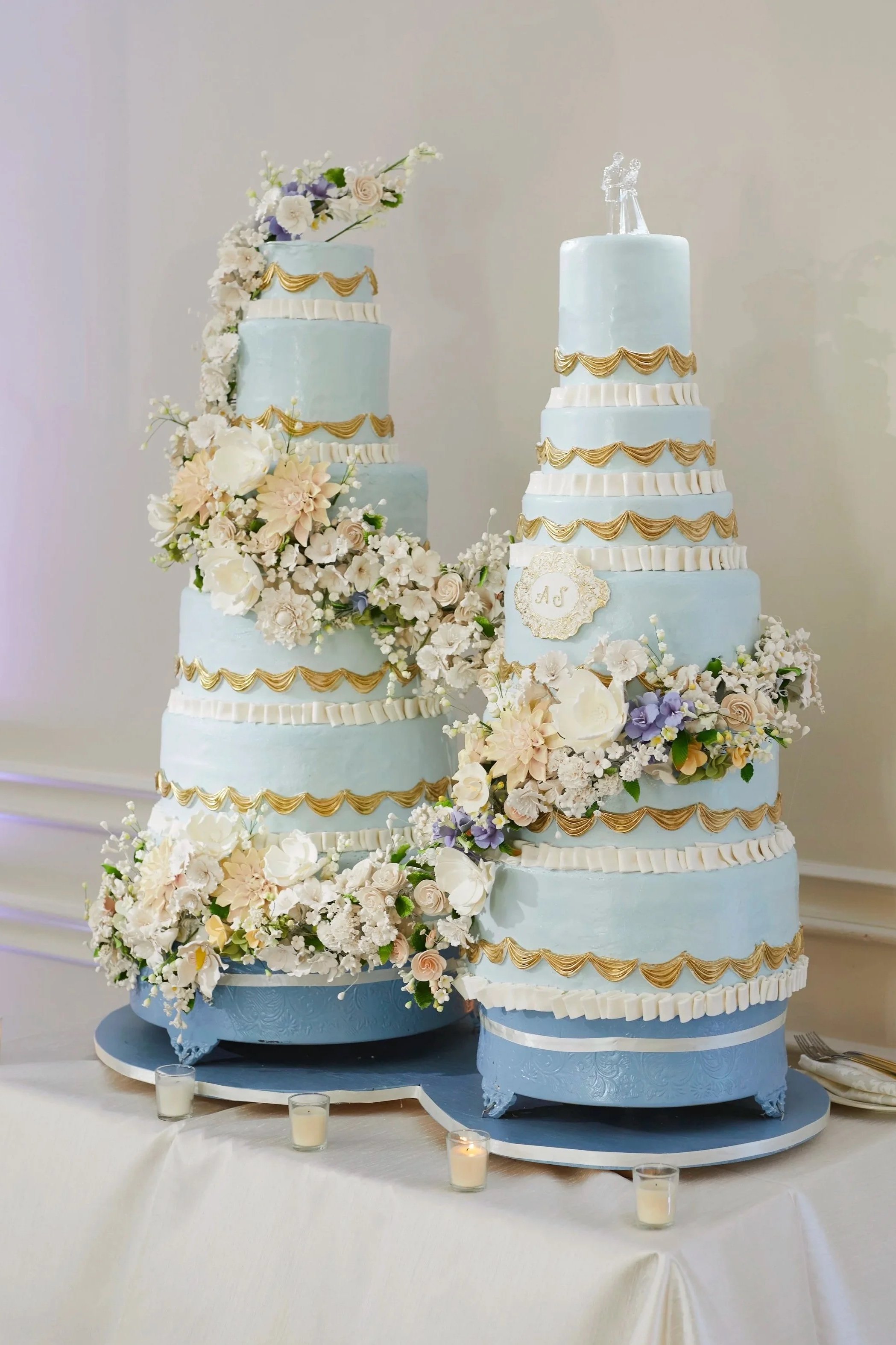 Samantha and Anthony’s custom wedding cakes designed by Executive Pastry Chef Daniel Andreotti,  featuring soft blue detailing, intricate sugar florals, and elegant gold accents displayed during the reception celebration. 