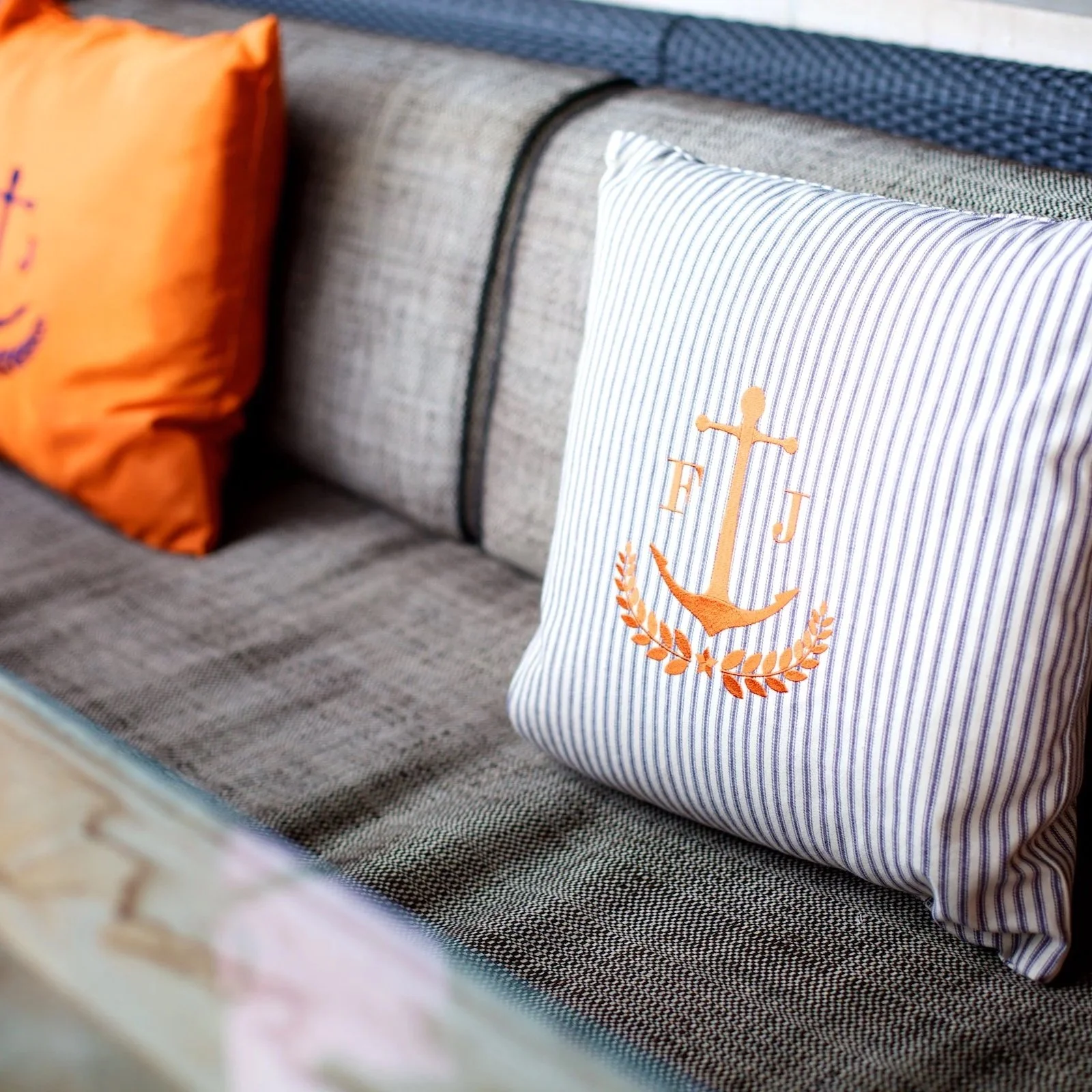 Close-up of decorative pillows on a grey upholstered sofa, with one white pillow featuring an embroidered orange anchor and letters 'F' and 'J' and laurel leaves, and others in orange and navy blue.