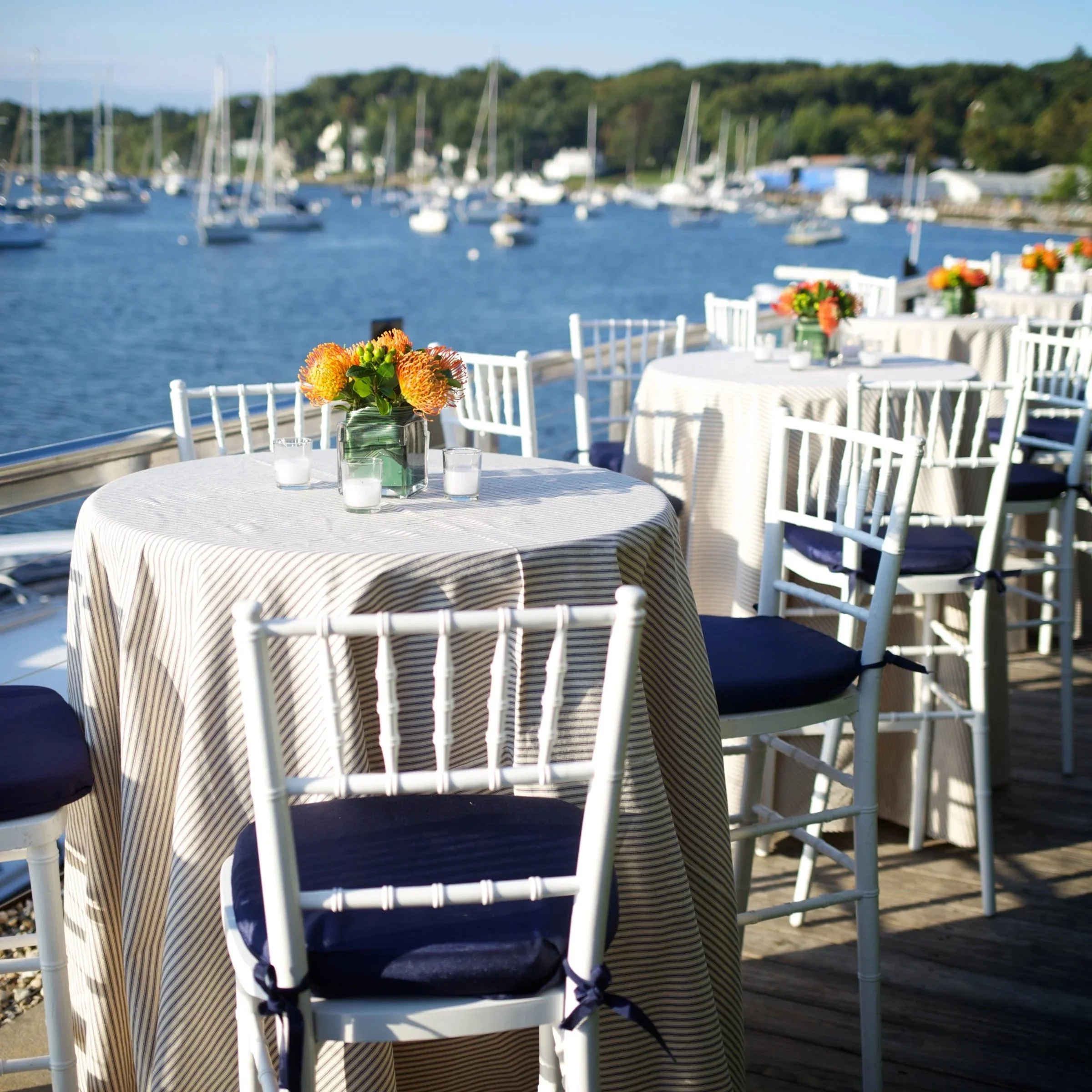 Outdoor dining setup on a deck by the water, with white chairs, tables with striped tablecloths, and floral centerpieces, boats docked in the marina in the background.