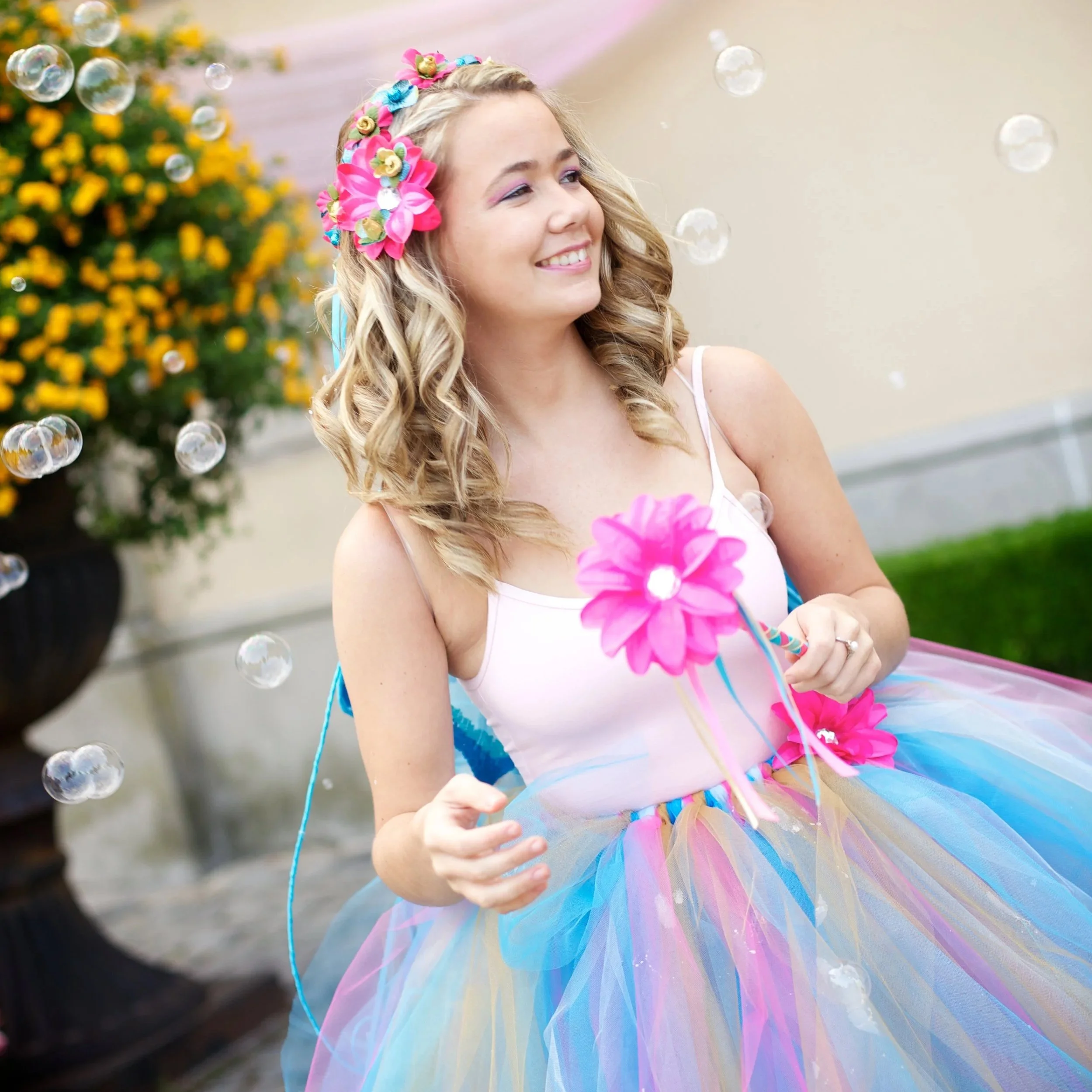 Young girl with long wavy blonde hair, wearing a pink outfit and a floral headband, smiling while holding a pink flower, surrounded by floating bubbles, with a colorful tutu skirt.