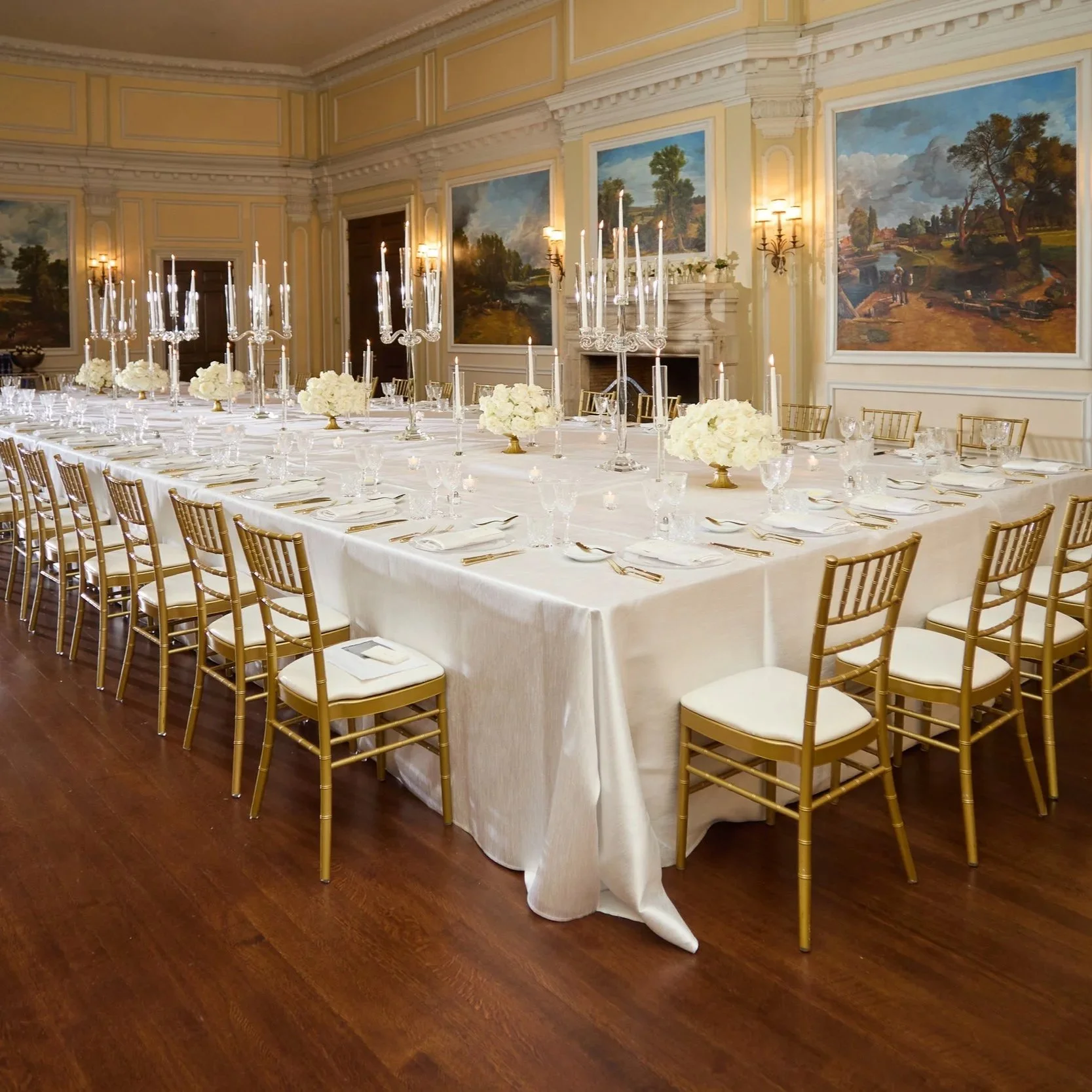 Elegant banquet hall with long rectangular table set for a formal event, decorated with white tablecloths, gold chairs, tall candelabras, floral centerpieces, and wall paintings of landscapes.