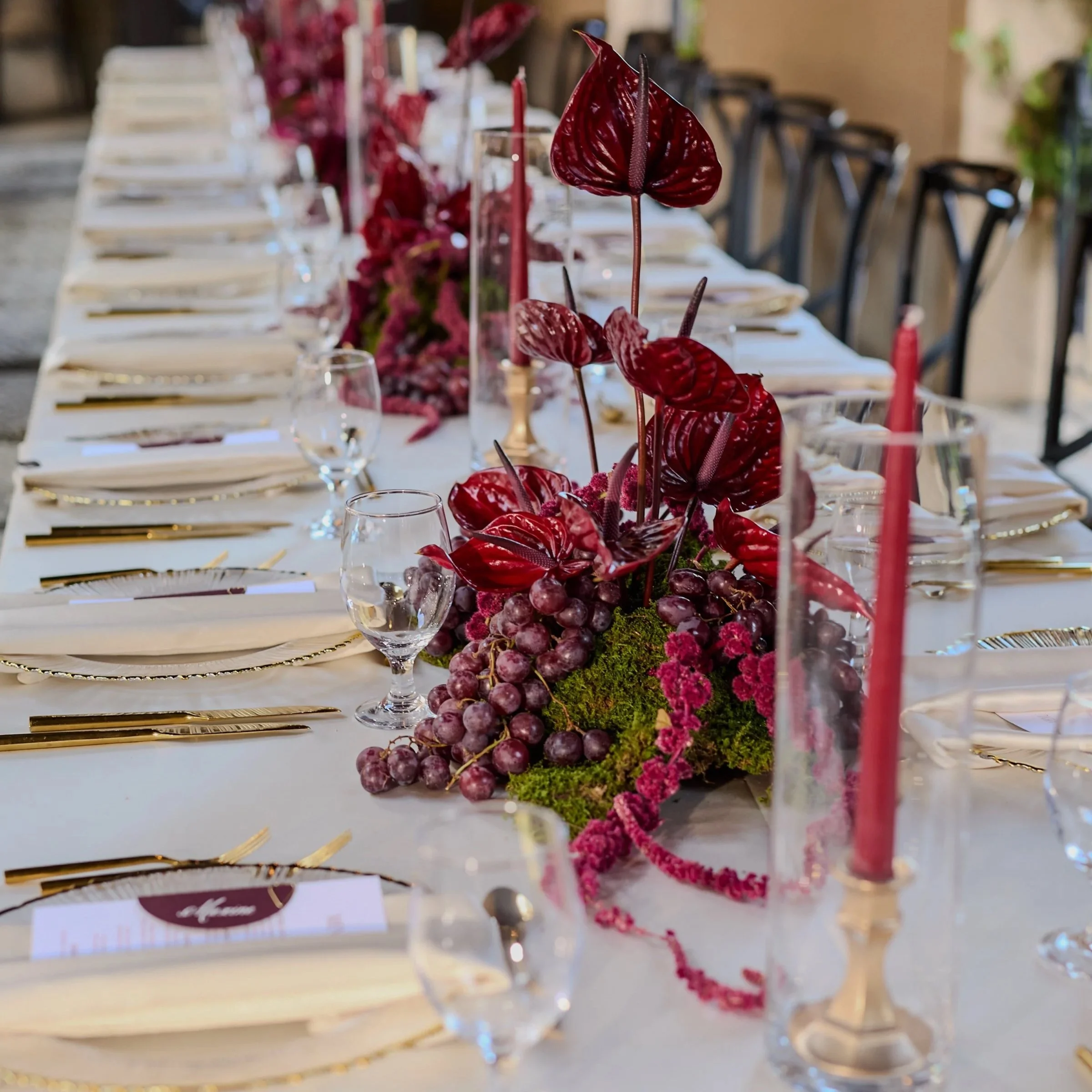 A long banquet table decorated with a floral centerpiece featuring dark red anthuriums and grapes, set for an event with plates, glasses, and gold utensils.