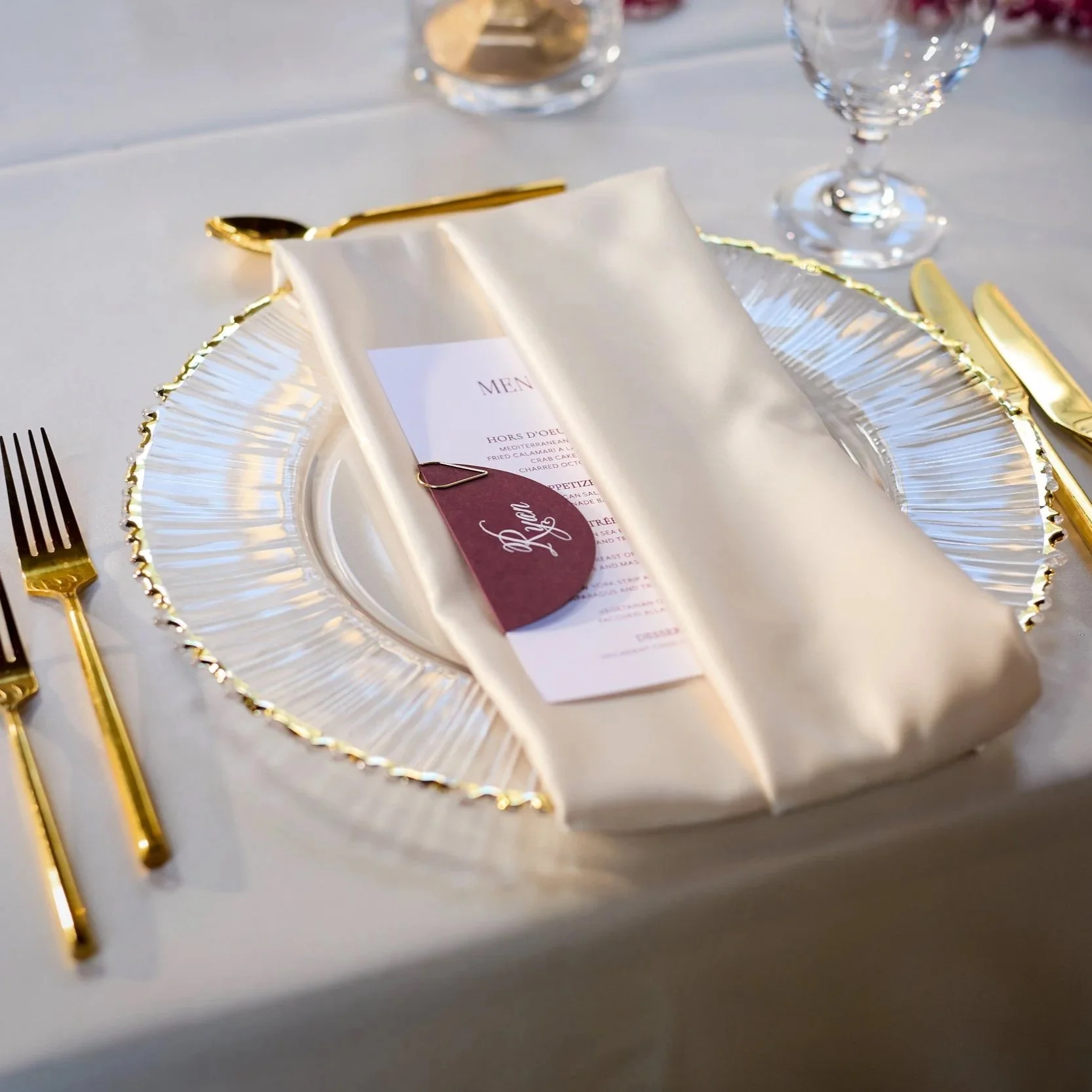 Elegant table setting with gold cutlery, a white napkin, and a menu card on a silver charger plate. Two glasses in the background, one of which is wine. A small looped tag labeled 'Dine' is placed on the menu.