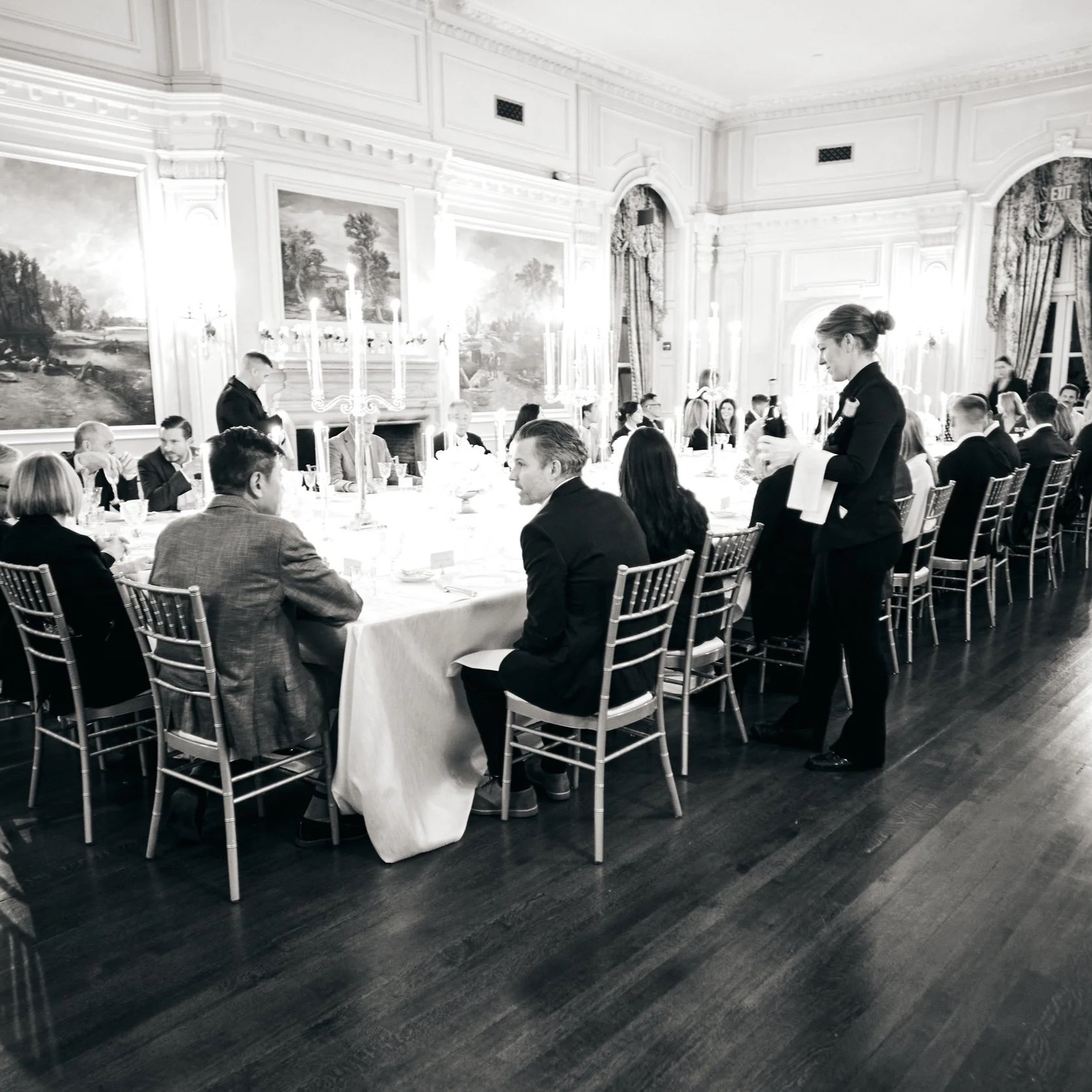 A formal dinner gathering in an elegant, historic-style room with painted landscape murals on the walls, ornate curtains, and wooden flooring. Multiple people are seated around a long table with a white tablecloth, and a woman in a black outfit is se