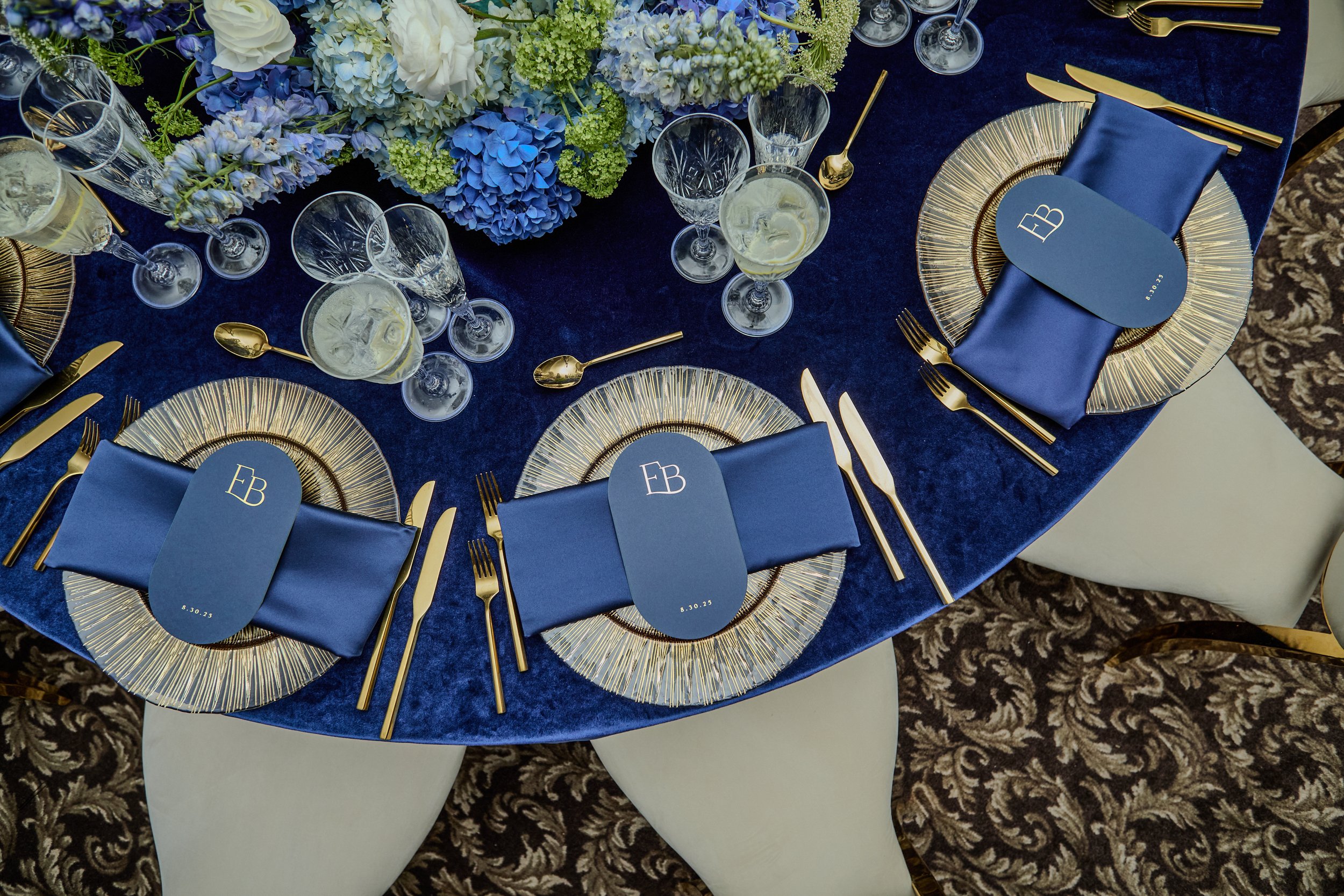 The reception unfolded in Oheka Castle’s Terrace Room, where king’s tables were designed in rich layers of deep blue, gold, and soft candlelight. Lush arrangements of hydrangea and delicate florals spilled across mirrored surfaces, creating a sense o