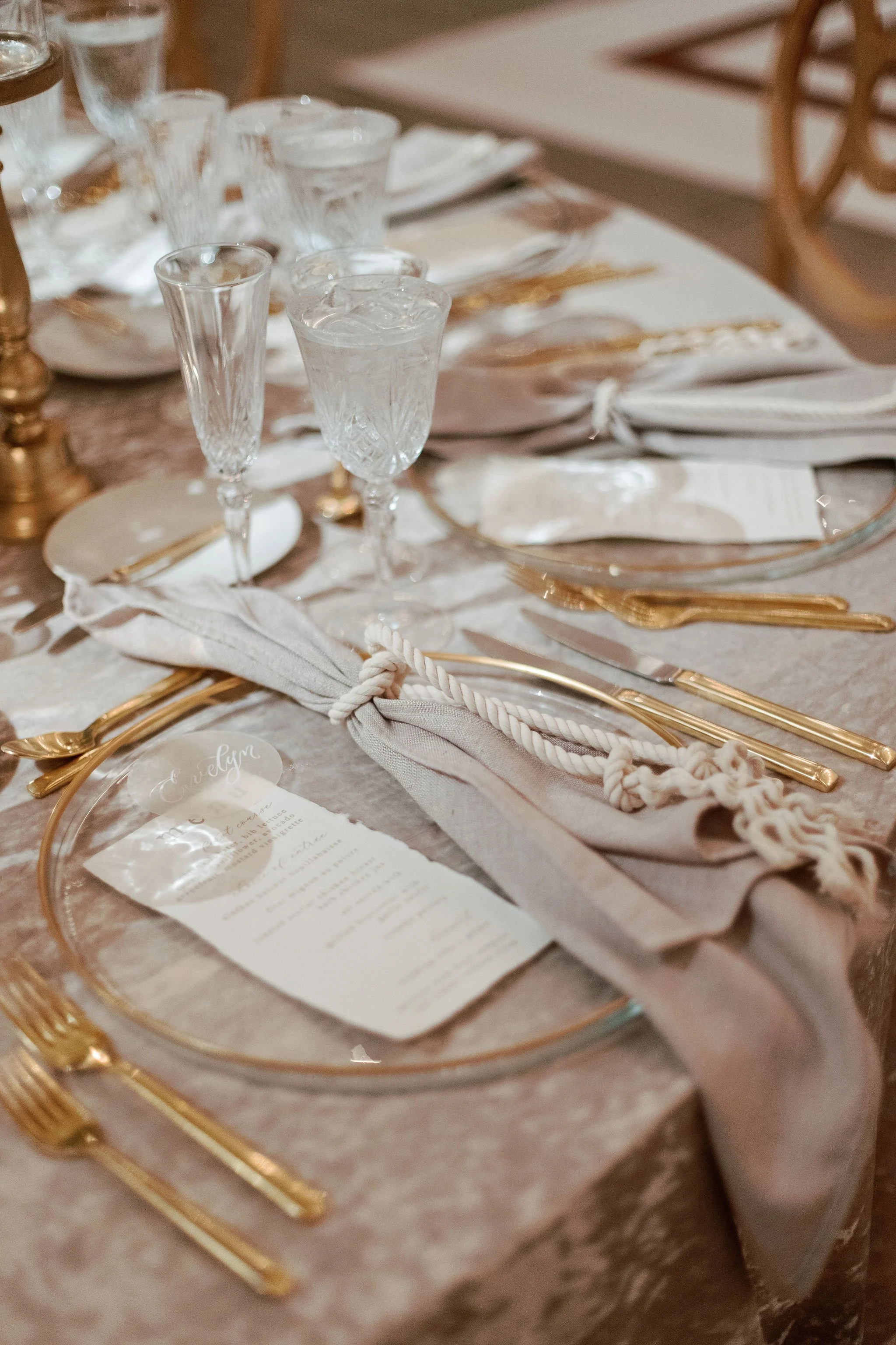A table setting with glassware, gold flatware, linen napkin, and a menu, arranged on a beige tablecloth.