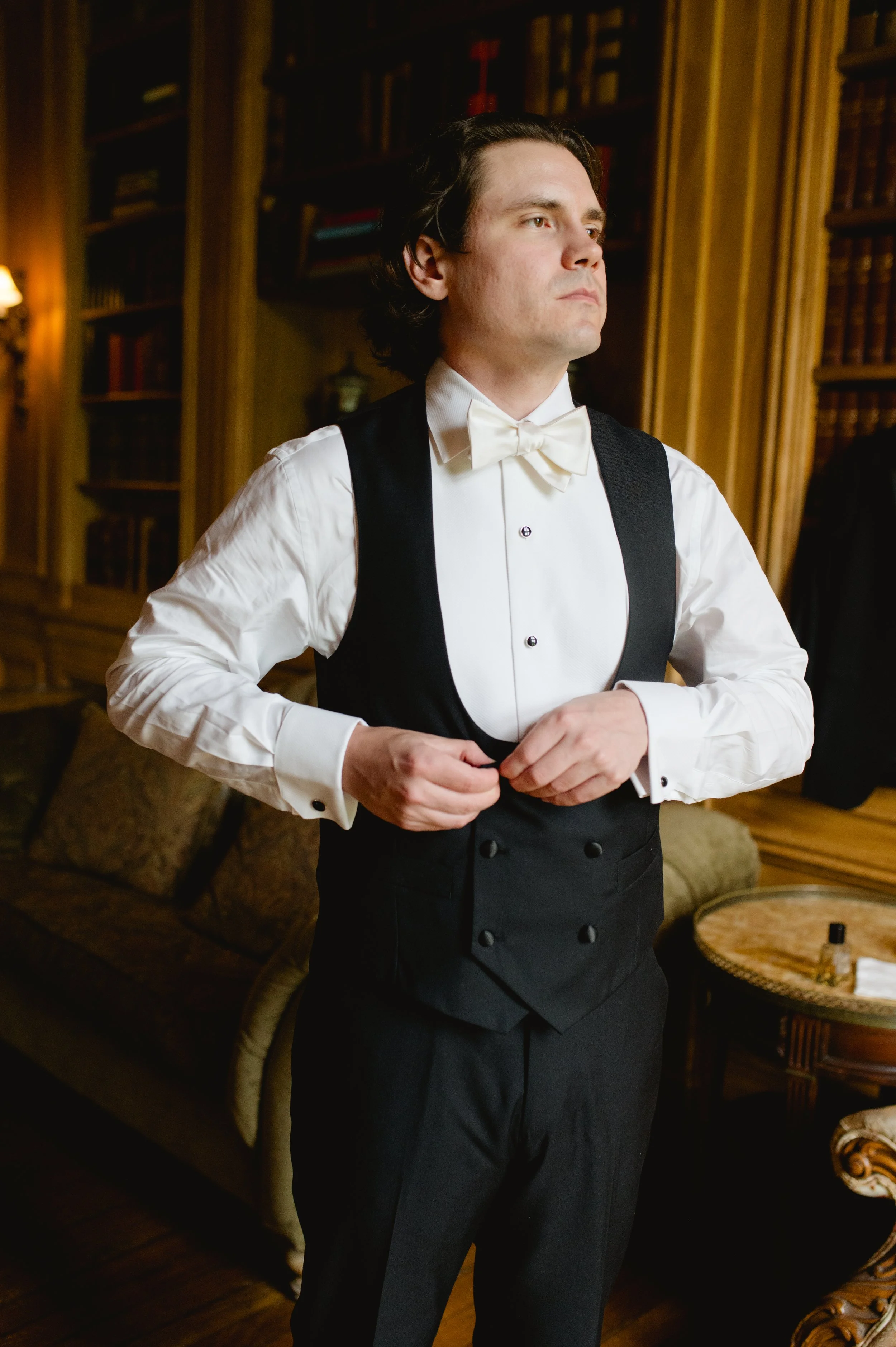 A man in formal attire, including a white shirt, black tuxedo vest, and bow tie, is adjusting his buttons in a room with wooden shelves and a sofa.