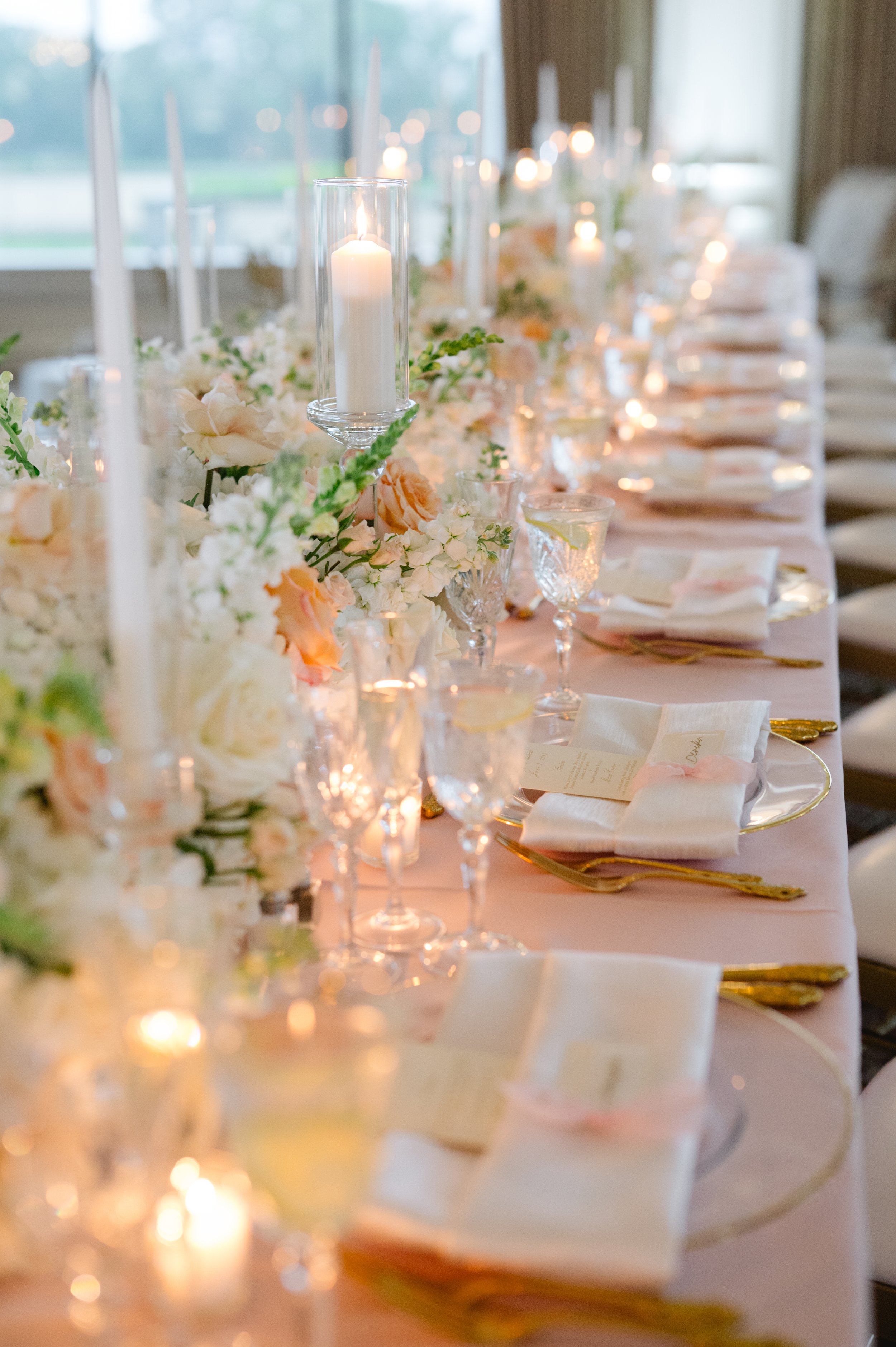 Elegant wedding reception table decorated with white and peach flowers, glass candles, and gold-rimmed glassware, set with white napkins and gold utensils.