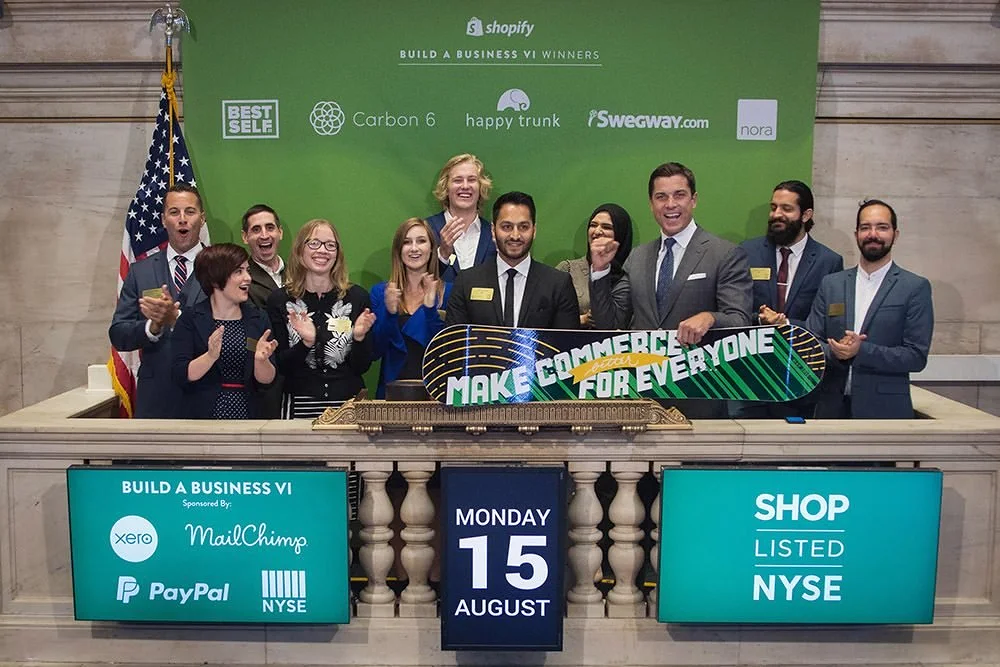 Group of people in business attire holding a skateboard with the message "Make Commerce for Everyone" in a formal setting, with a green backdrop displaying company logos. The date is August 15, and they are celebrating the Build a Business VI event sponsored by various companies.