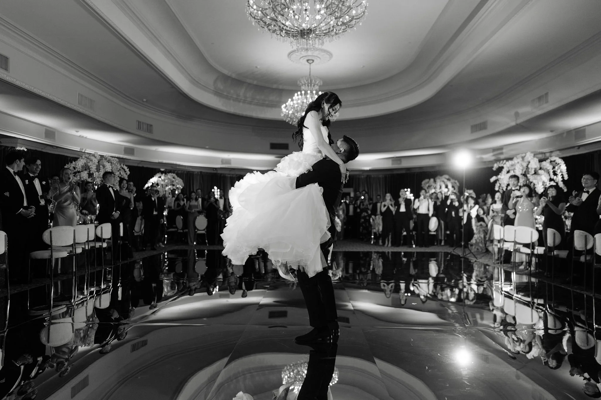 A wedding reception with a bride being lifted by the groom on a dance floor, surrounded by guests in formal attire, and decorated with floral arrangements and chandeliers.