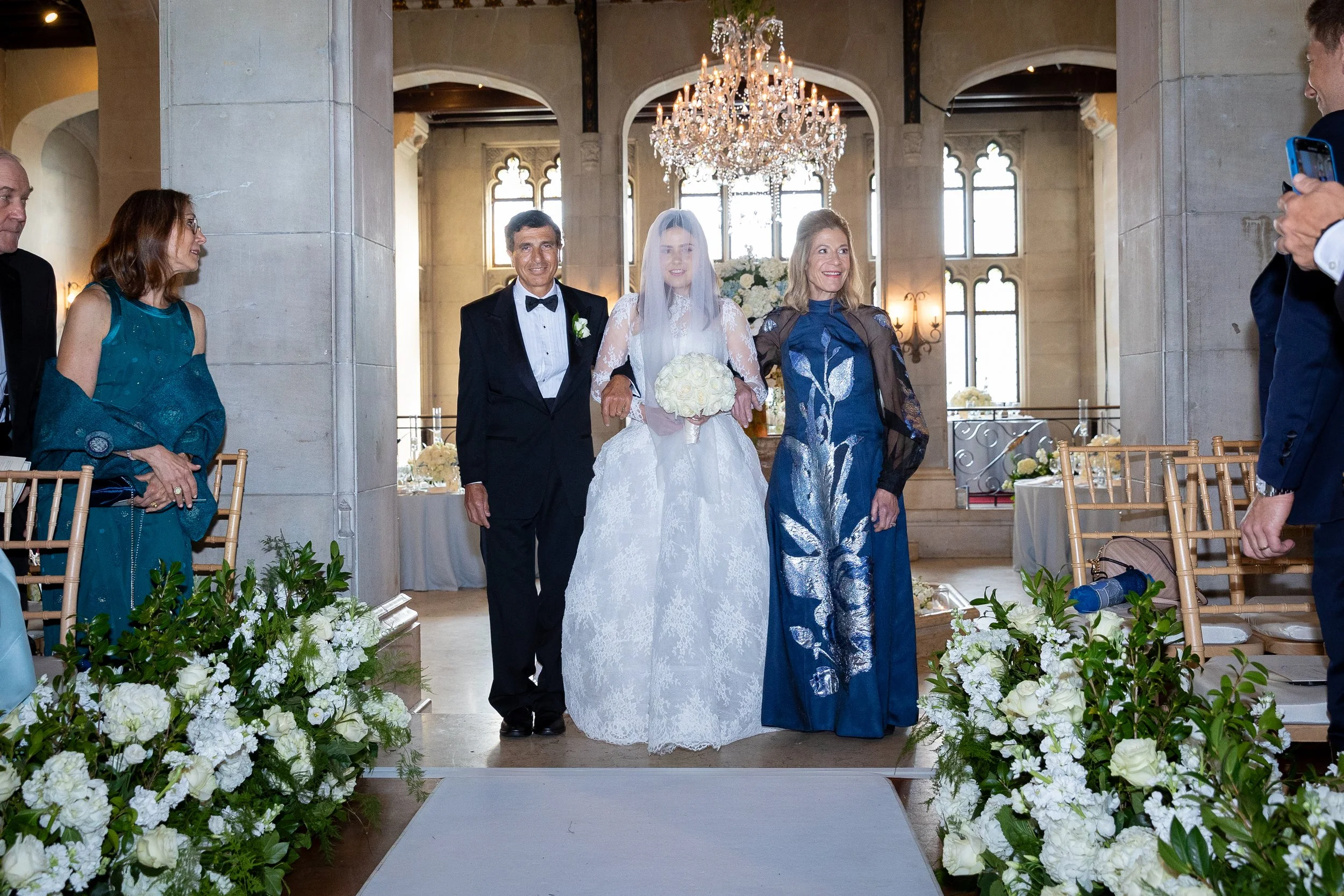 Bride in a white lace dress and veil holding a bouquet walking down the aisle with her parents during a wedding ceremony in a decorated hall with floral arrangements and a chandelier overhead.