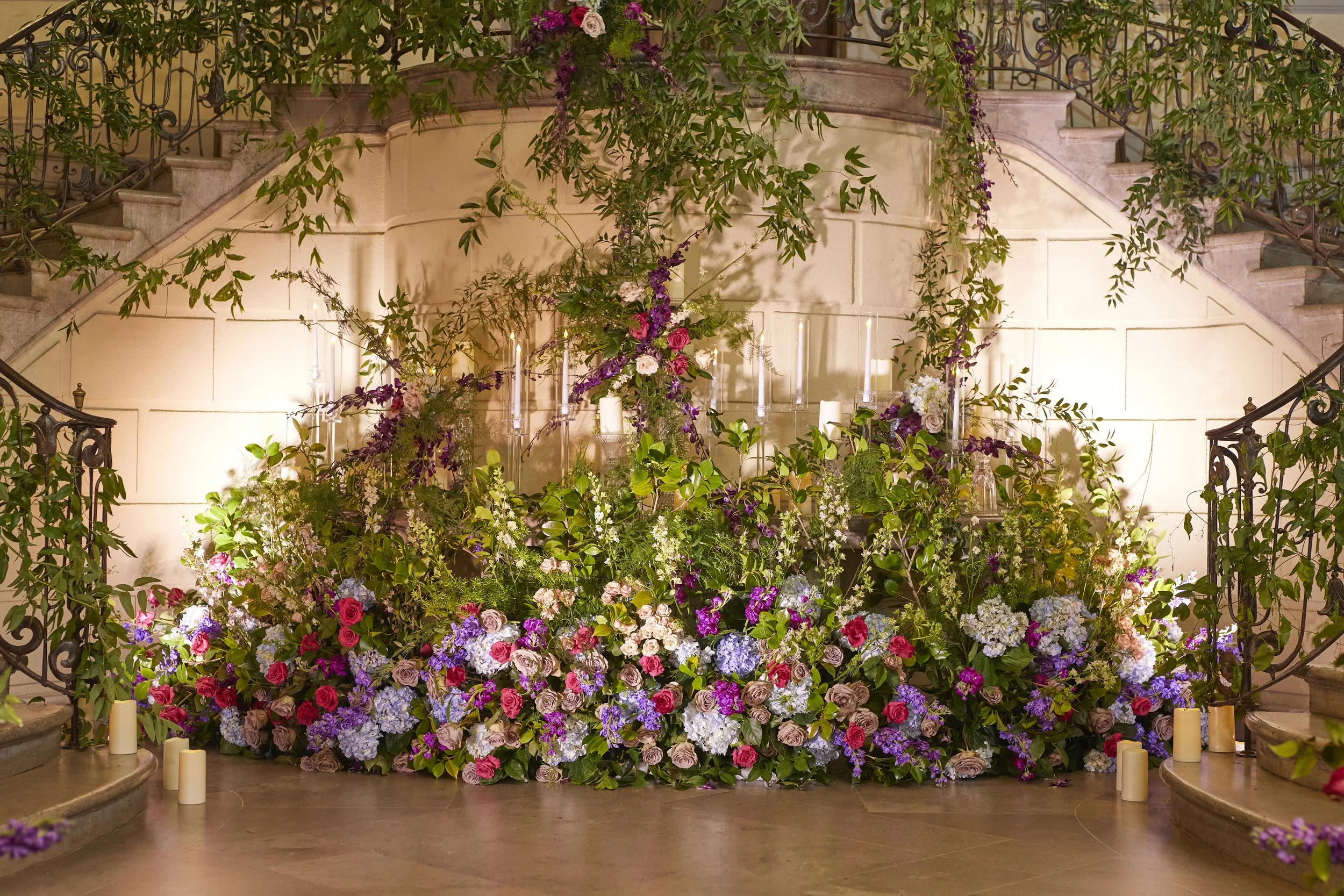 A romantic floral installation layered along the grand staircase at Oheka Castle with lush blooms and candlelight creating an immersive reception backdrop.