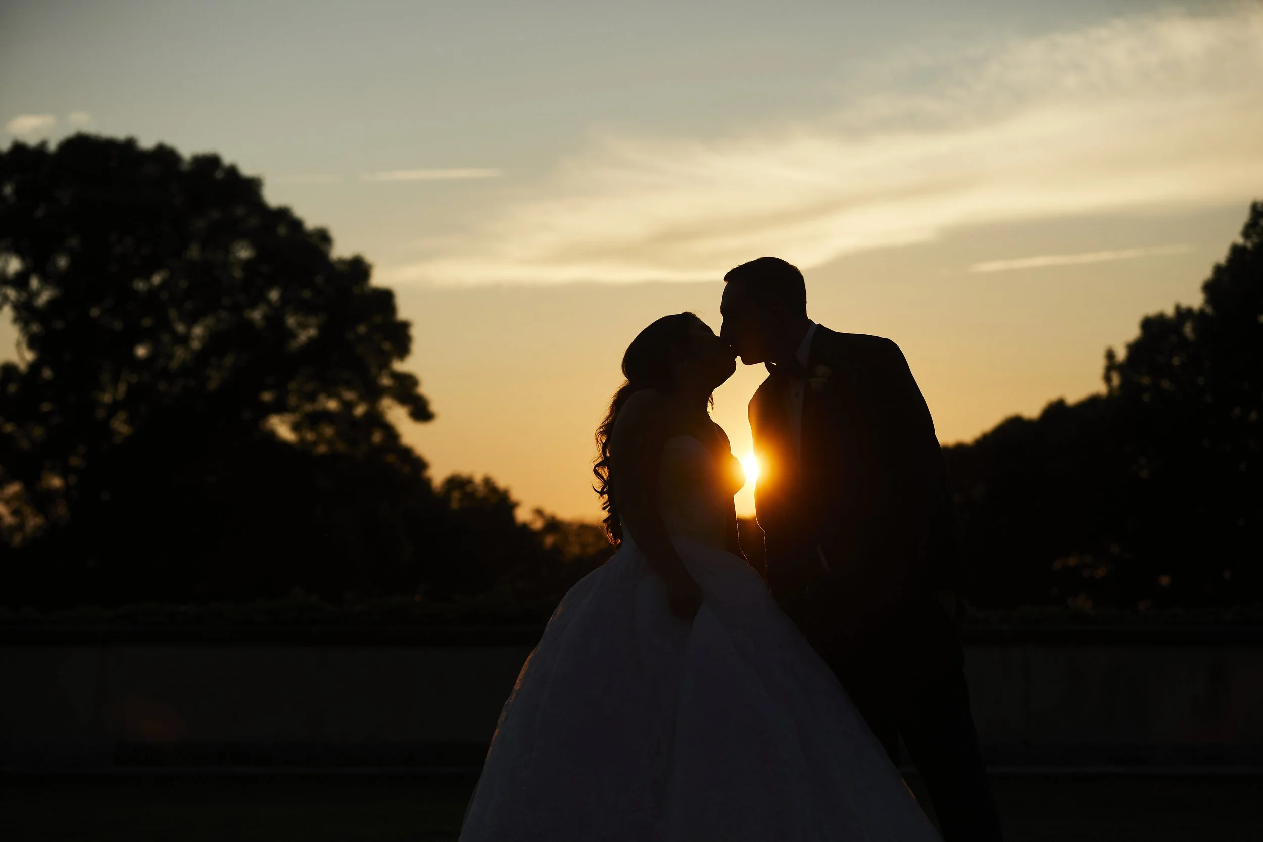 Samantha and Anthony sharing a romantic sunset kiss silhouetted against the evening sky on the grounds of Oheka Castle.