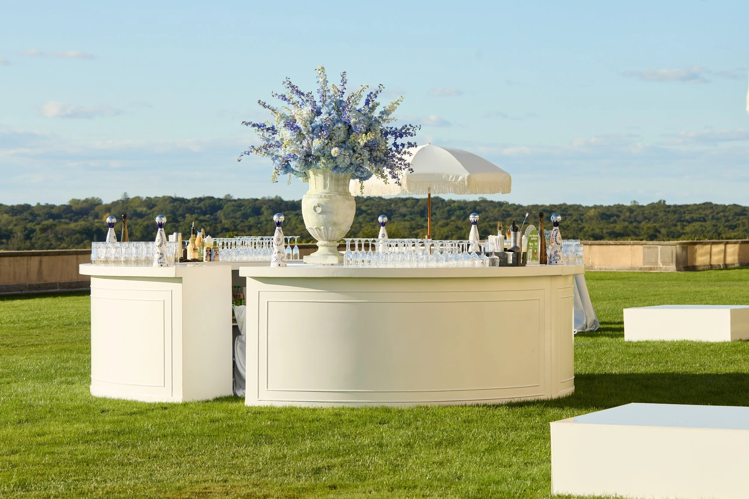 On the great lawn, a custom-designed circular bar anchored cocktail hour, styled in soft ivory tones and topped with an abundant arrangement of blue and white florals. Set against sweeping views of the estate, the space felt open, elegant, and though