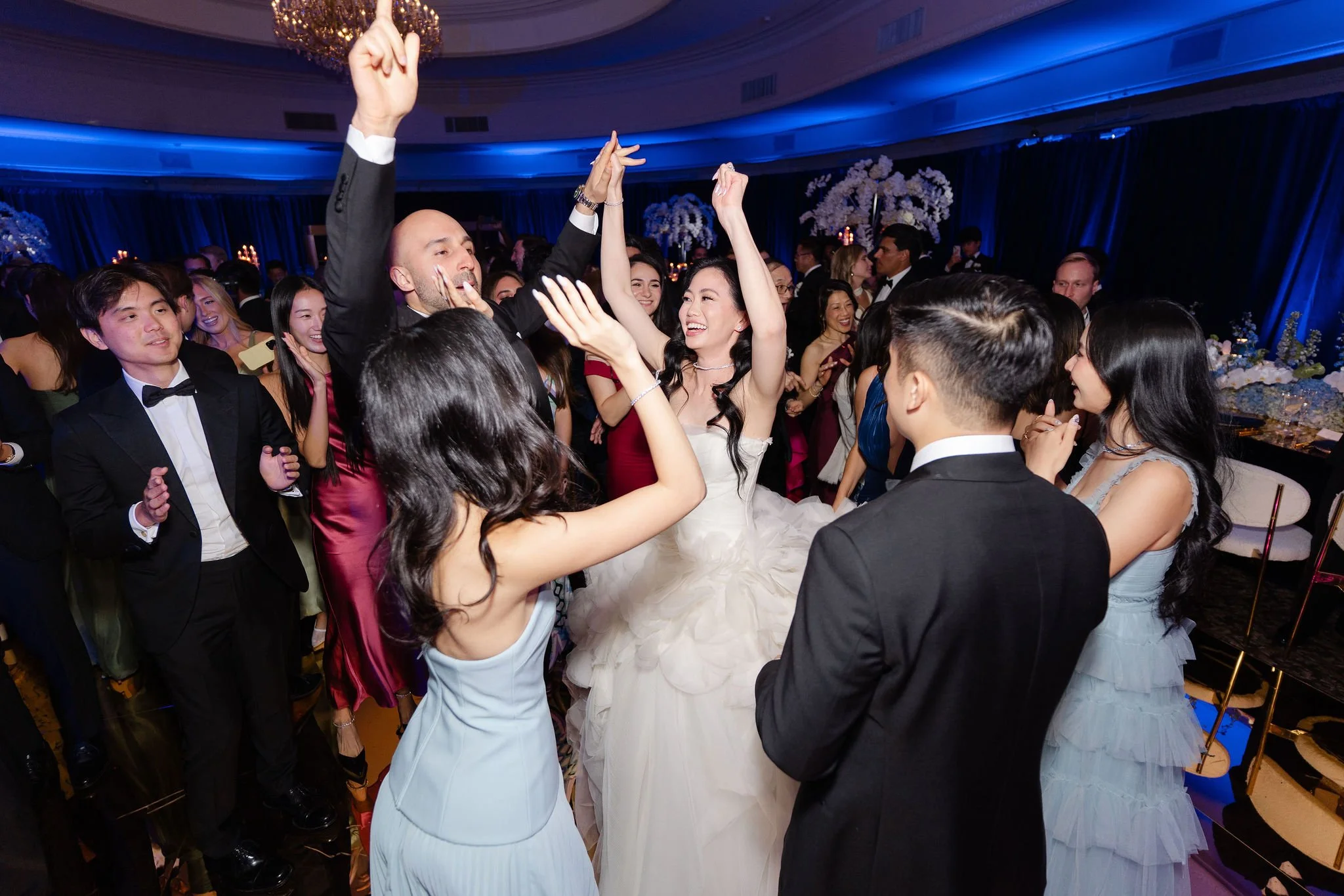 Group of people dancing and celebrating at a wedding reception, with a bride in a white dress and a groom among the guests.