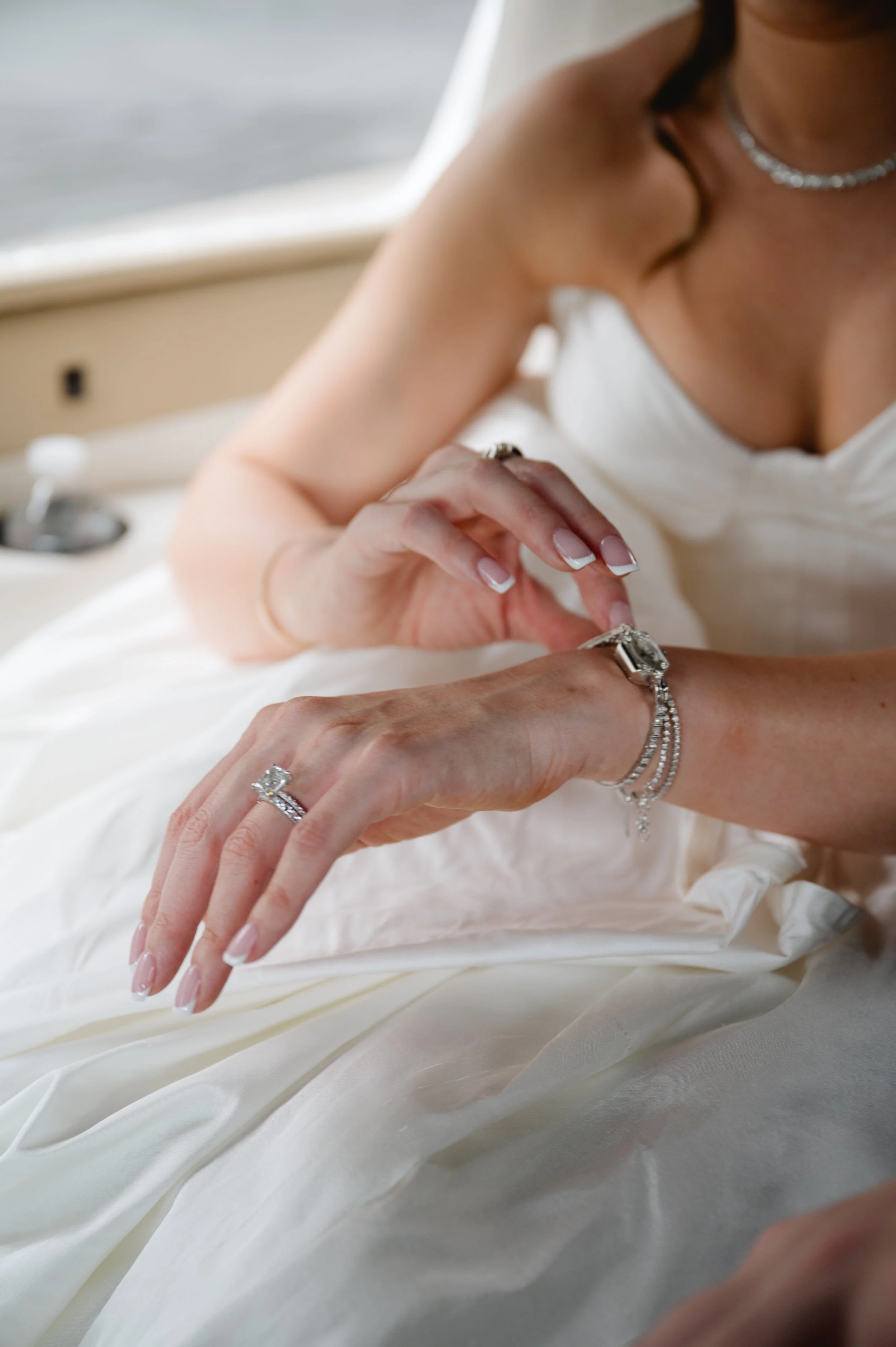 Close-up of a bride with light skin adjusting her diamond ring on her finger, wearing a white wedding dress, jewelry including a ring, a bracelet, and a necklace, on a boat or yacht with a window in the background.