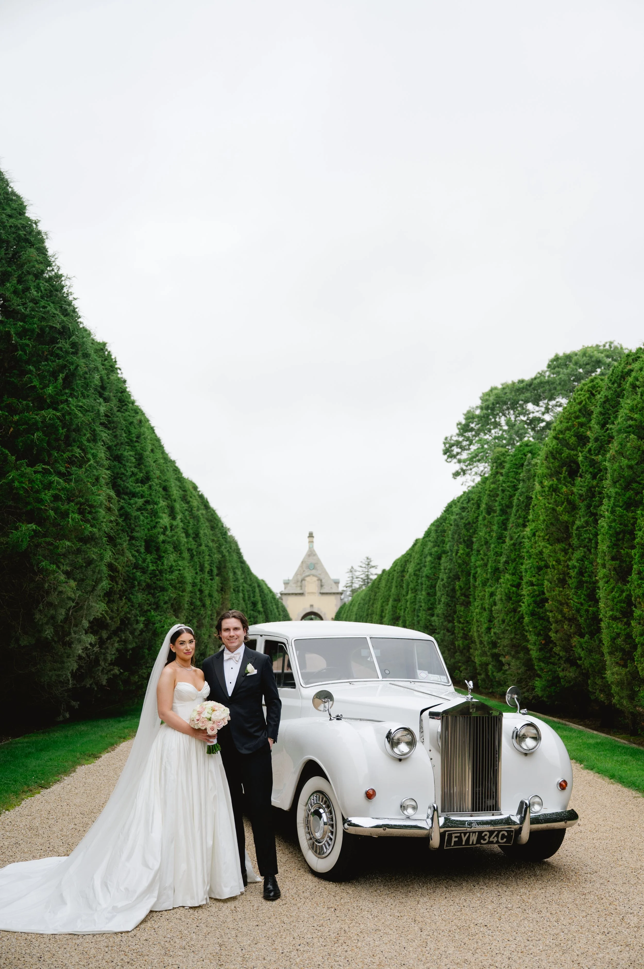 A bride and groom standing beside a classic white Rolls Royce on a gravel pathway, with manicured green trees and a historic building with a tower in the background.