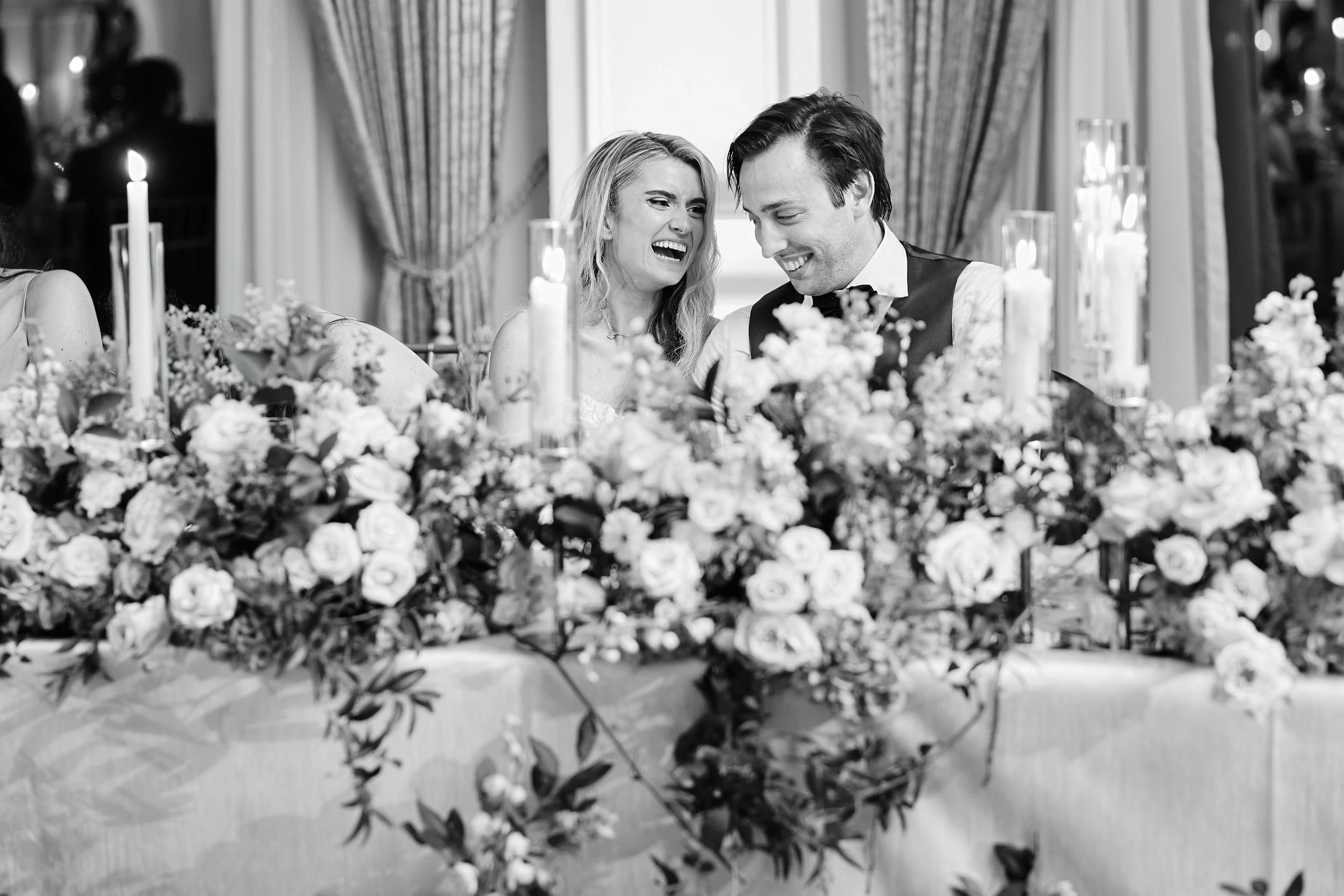 A bride and groom sharing a joyful moment at their wedding reception, sitting at a table decorated with flowers and candles.