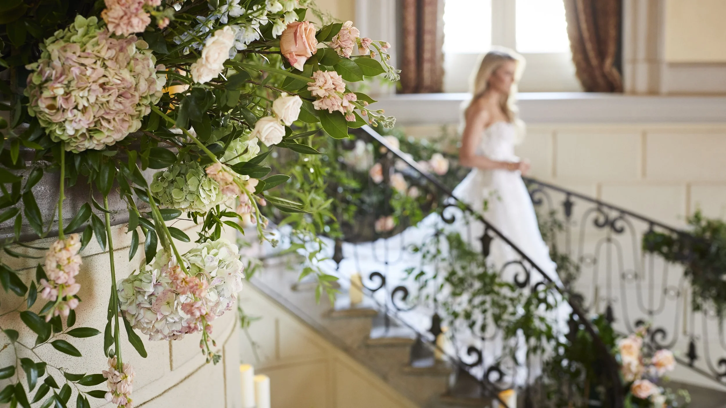 Pink and white hydrangea flowers with green leaves in the foreground, blurred woman in a white dress standing on a staircase in the background.