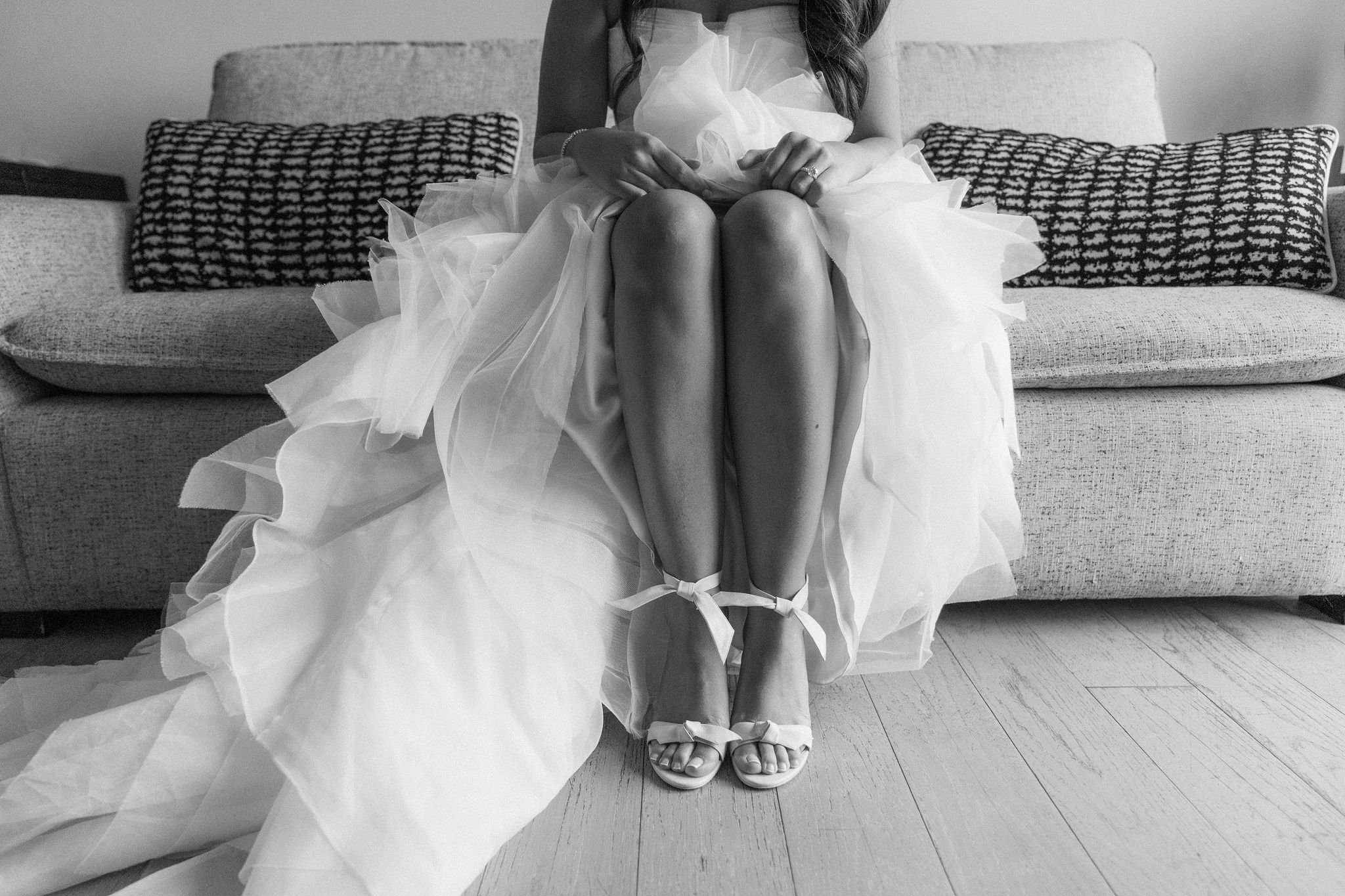 Black and white photo of a woman sitting on a sofa with her knees close together. She is wearing a strapless dress with a large tulle skirt and ballet slippers, with a ribbon tied around her ankles. Her hands are resting on her knees, and she is wear