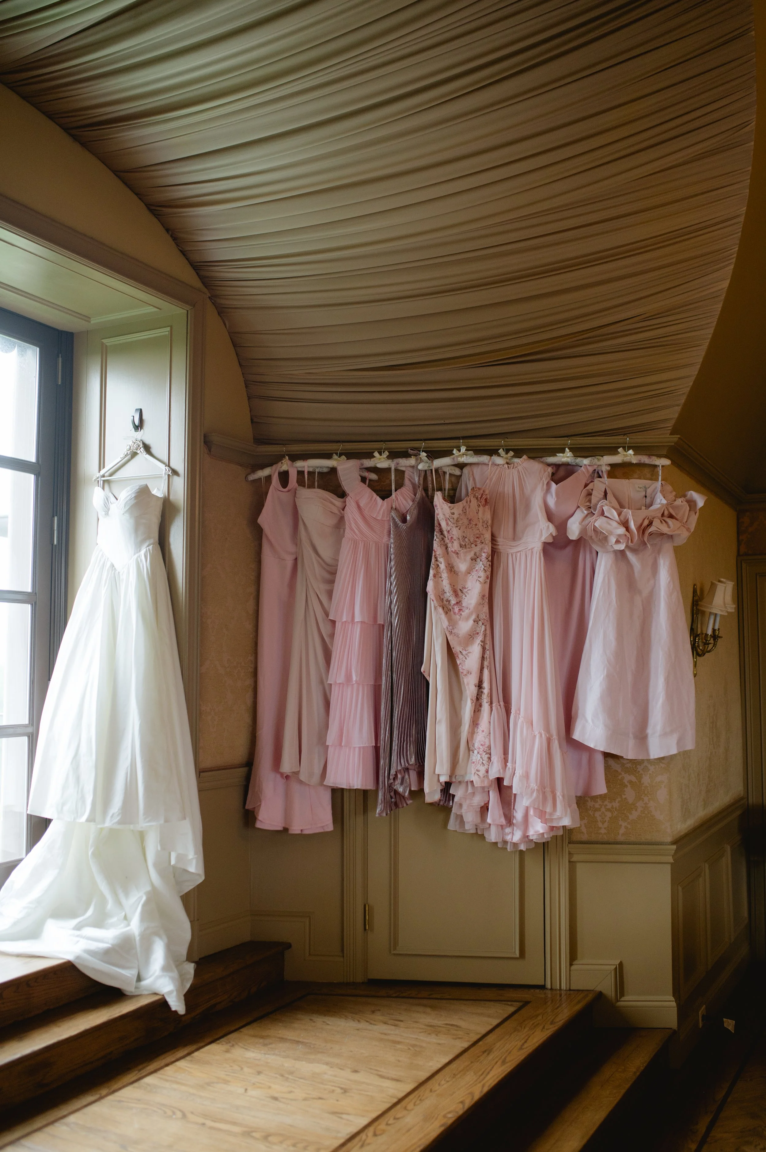 A row of pink and floral dresses hanging on a rack near a window, with a white wedding gown on a hanger on a nearby wall in a room with wooden stairs and ornate wall detailing.