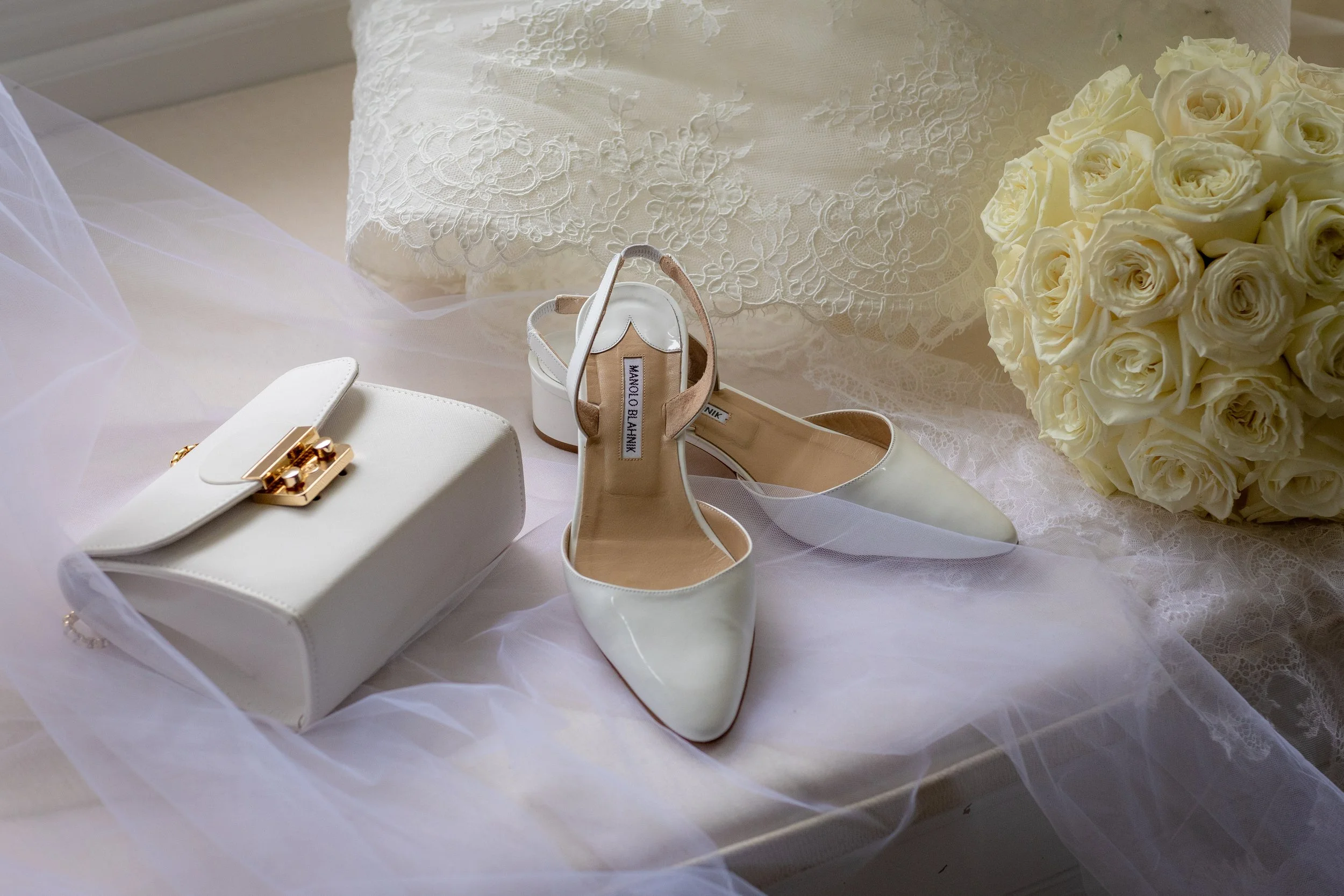 A collection of white wedding accessories including a bouquet of white roses, bridal shoes, a purse, and lace fabric.