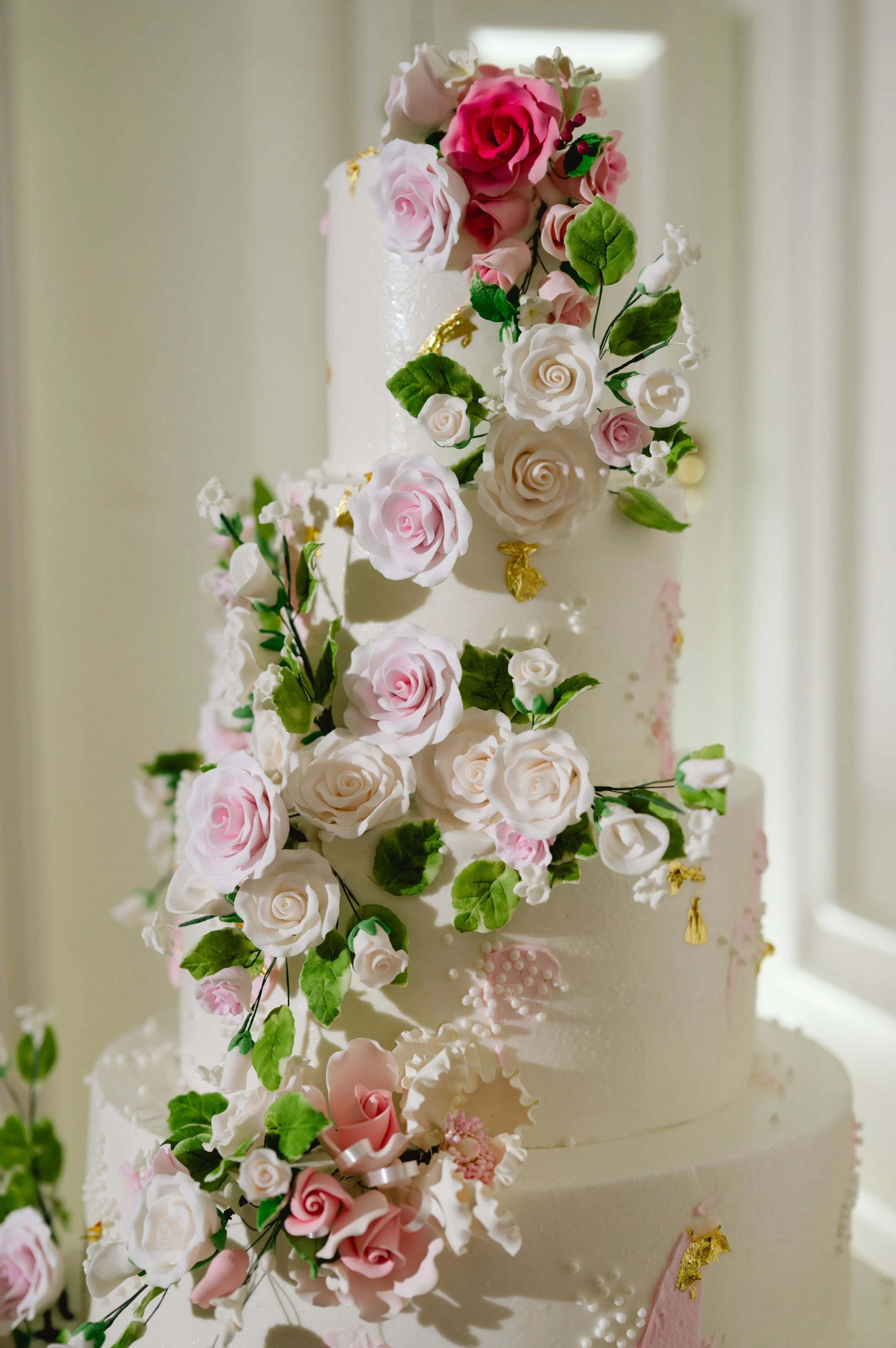 Elegant wedding cake decorated with cascading pink and white roses, green leaves, and gold accents.