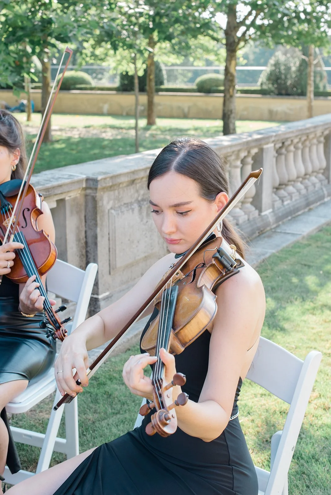 A young woman playing a violin outdoors during daytime, with another violinist partially visible beside her, in a park-like setting with trees and a stone wall in the background.