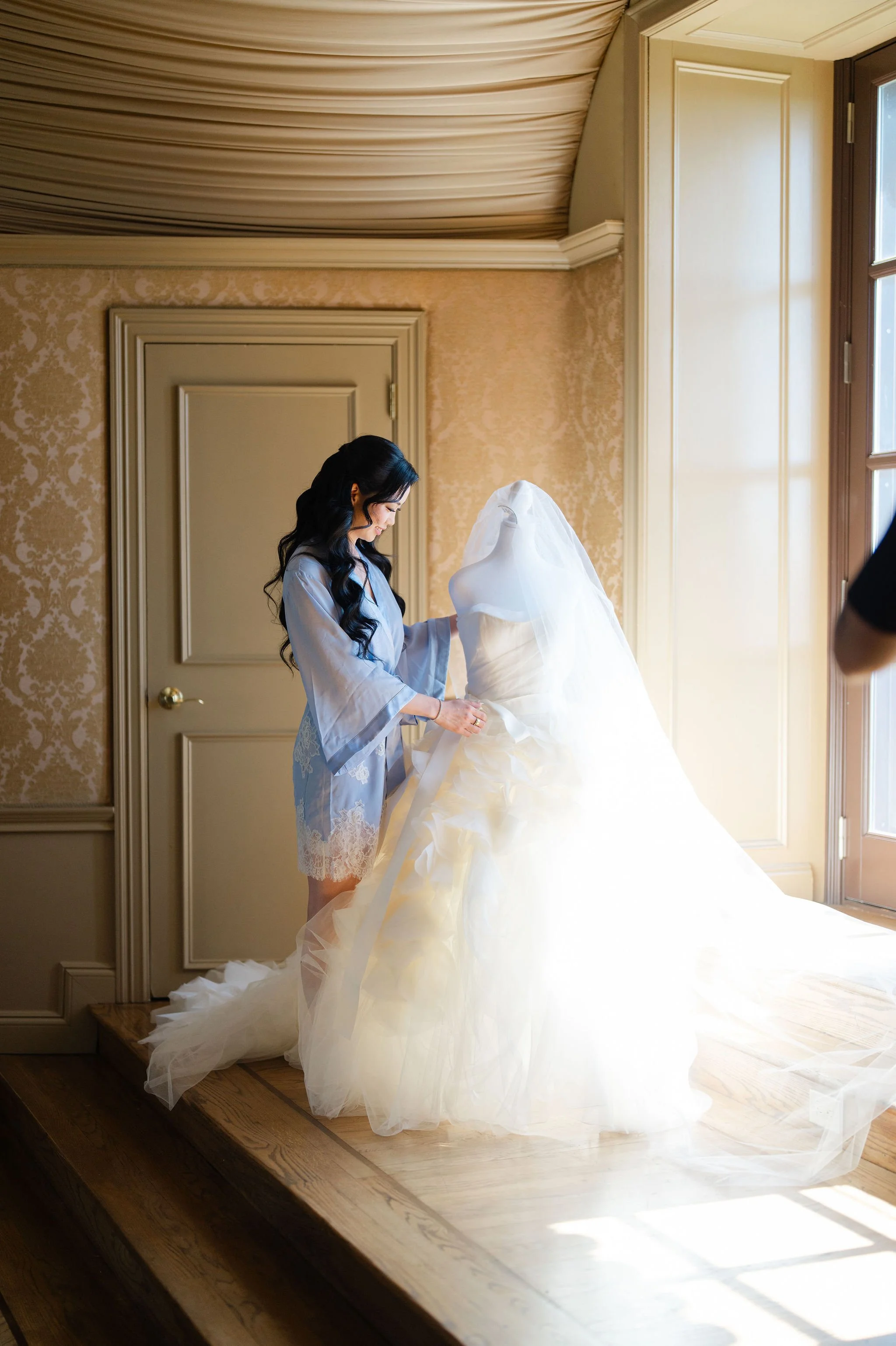 A woman in blue pajamas is standing on a wooden platform, holding and arranging a wedding dress, while bright natural light streams through a large window in a warmly decorated room.