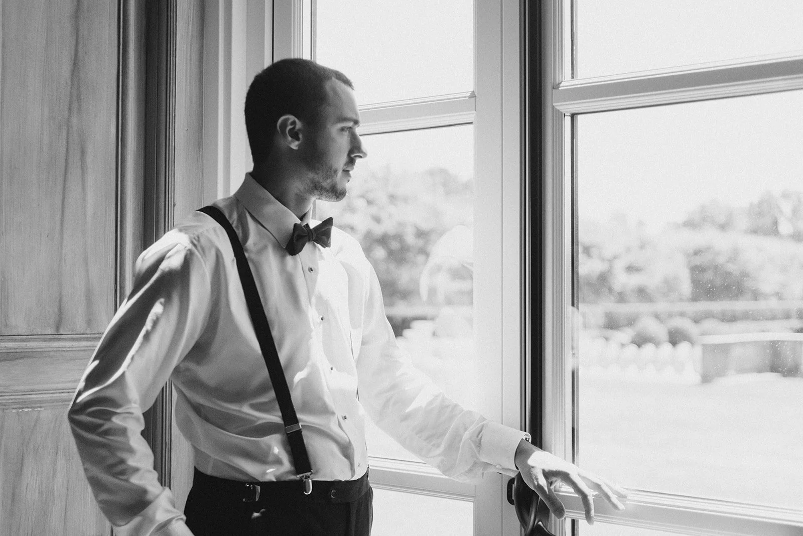 A man dressed in formal attire with a bow tie and suspenders, standing by a window and looking outside in black and white.