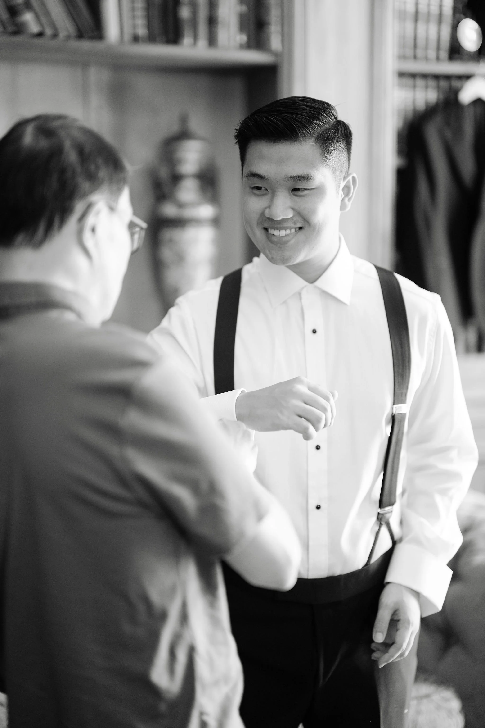 A young man in a white shirt and suspenders smiling and shaking hands with an older man in a suit inside a room with bookshelves and a decorative vase.