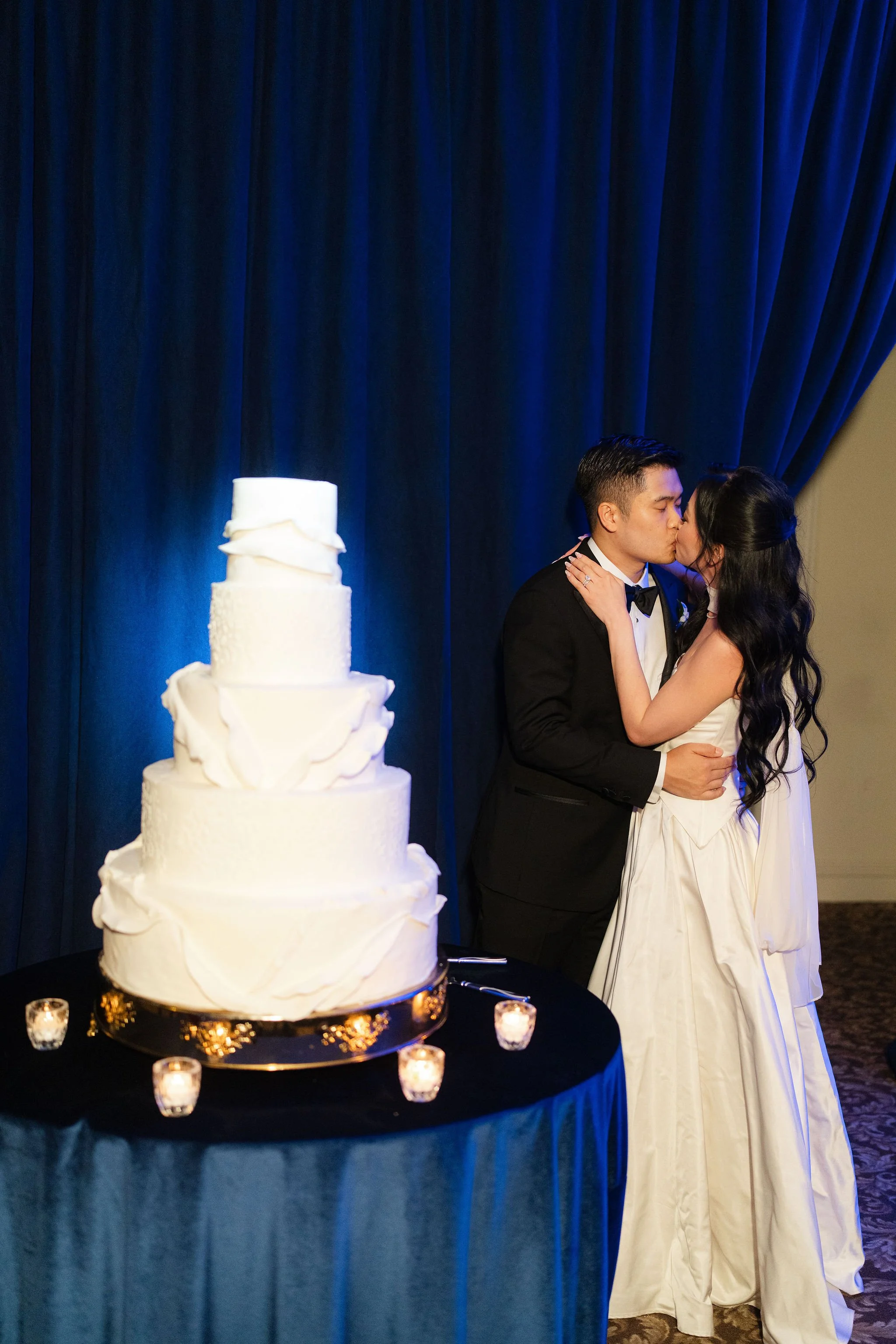 A wedding couple sharing a kiss next to a tiered white wedding cake decorated with ruffles, set on a table with small lit candles, against a dark blue curtain backdrop.