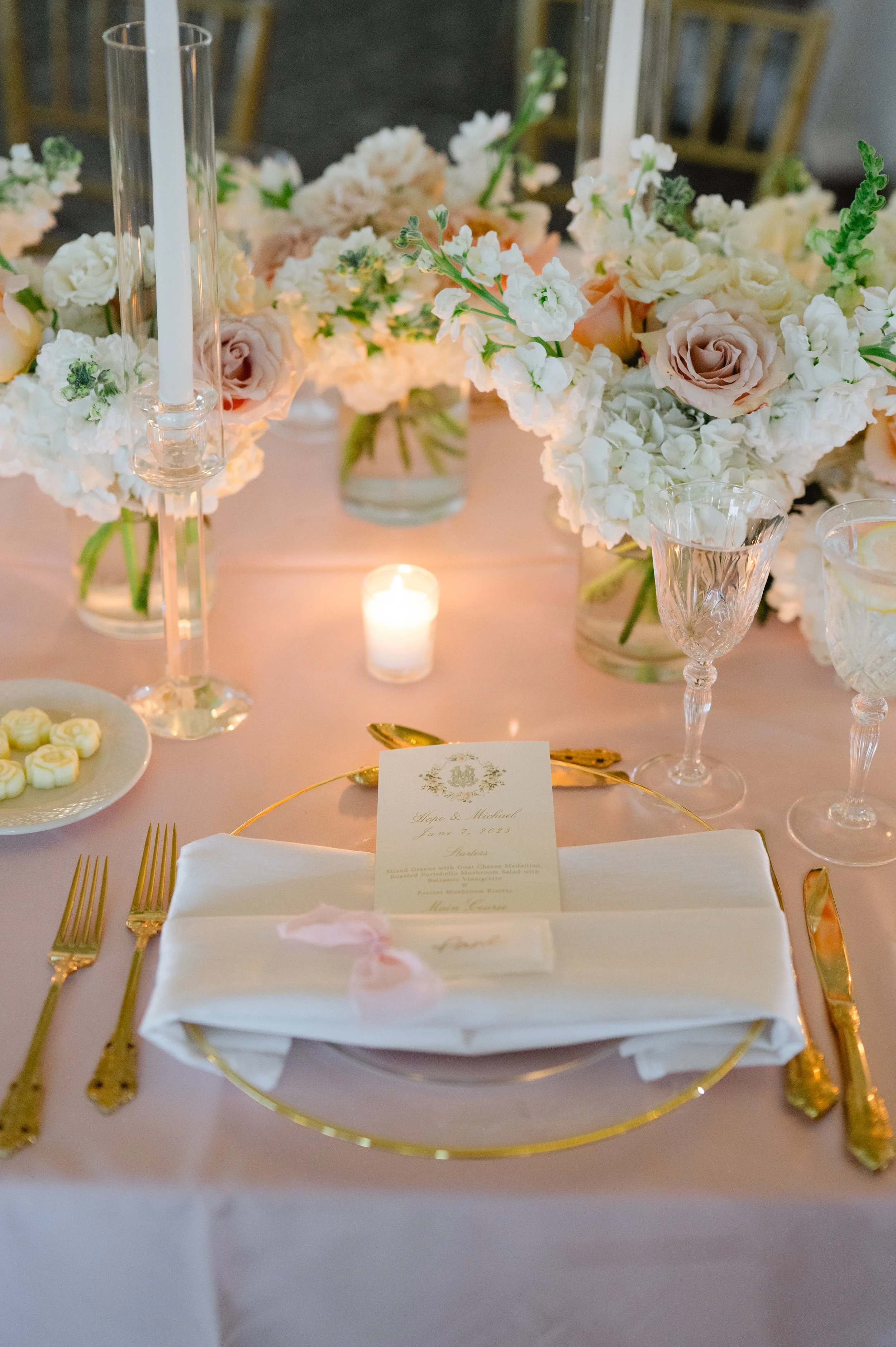Elegant wedding reception table setting with white and blush floral centerpieces, gold flatware, a white napkin with a pink ribbon, a wedding menu card, and lit candles.