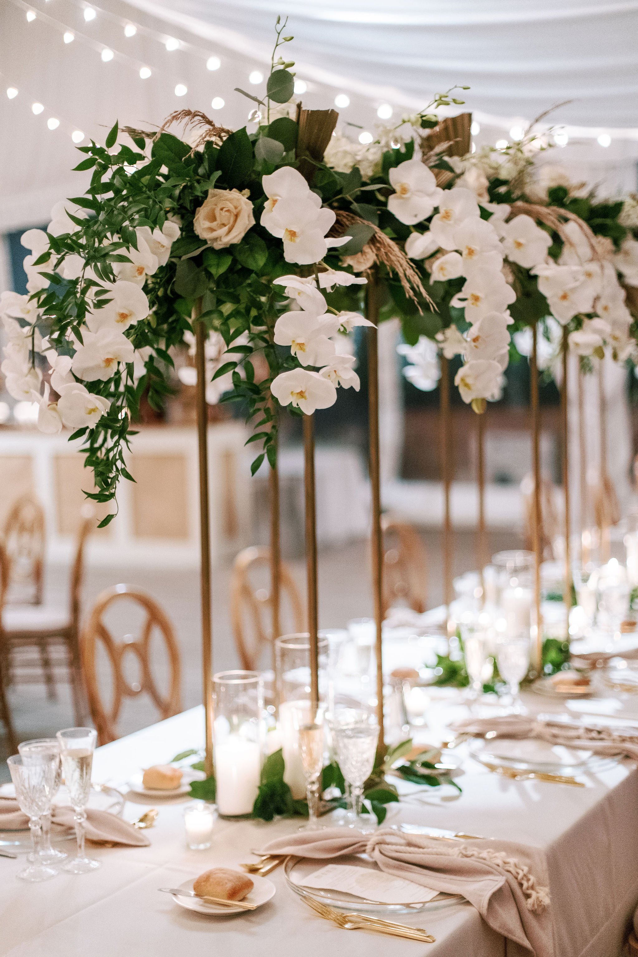 A table at a wedding reception decorated with a tall floral centerpiece featuring white orchids, roses, greenery, and wooden accents. The table is set with glassware, napkins, bread rolls, and plates, with chairs arranged around it. Background has st