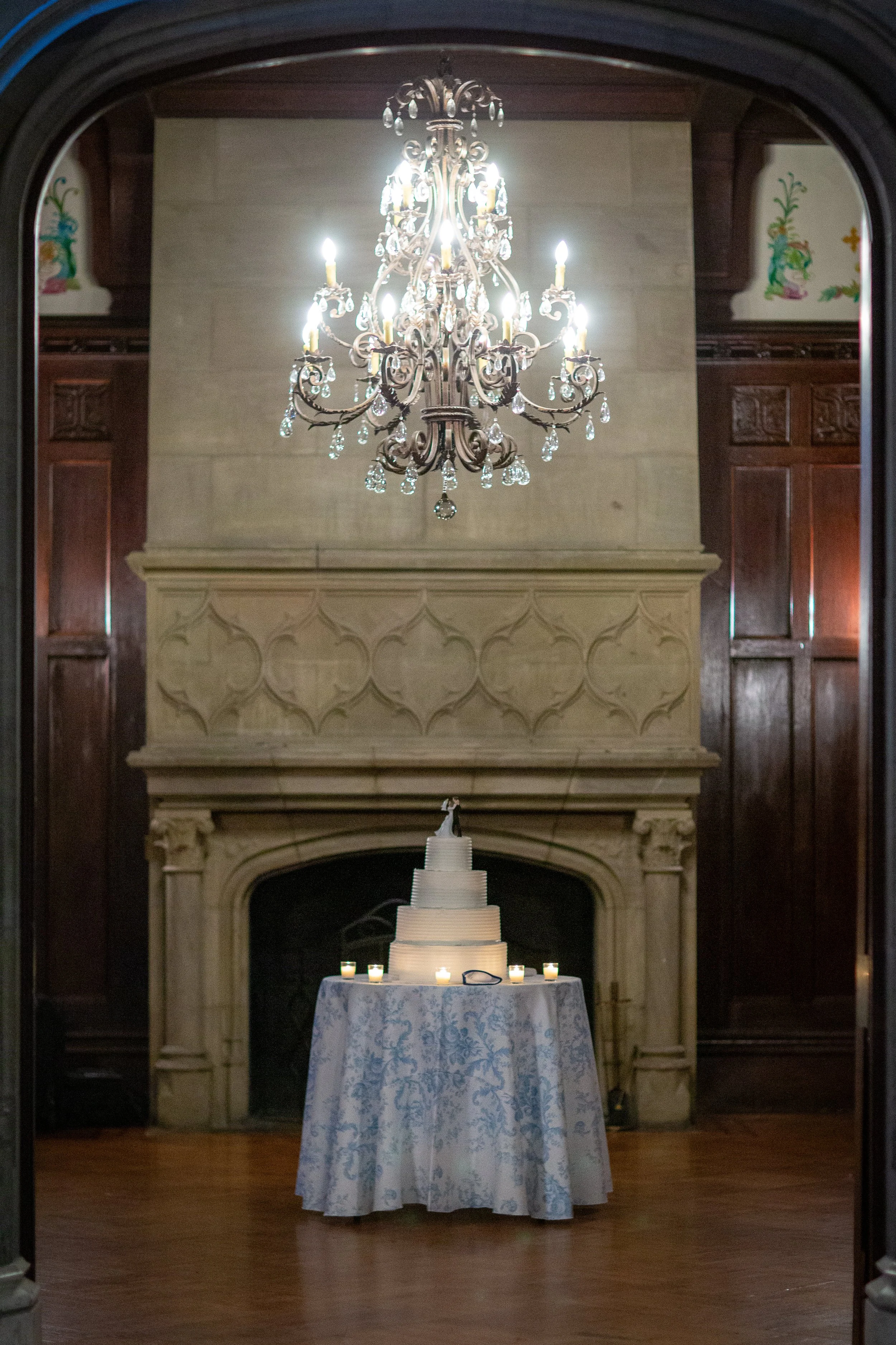 Chandelier hanging from the ceiling, illuminating a fireplace with decorations on top, positioned behind a table covered with a white tablecloth and candles.