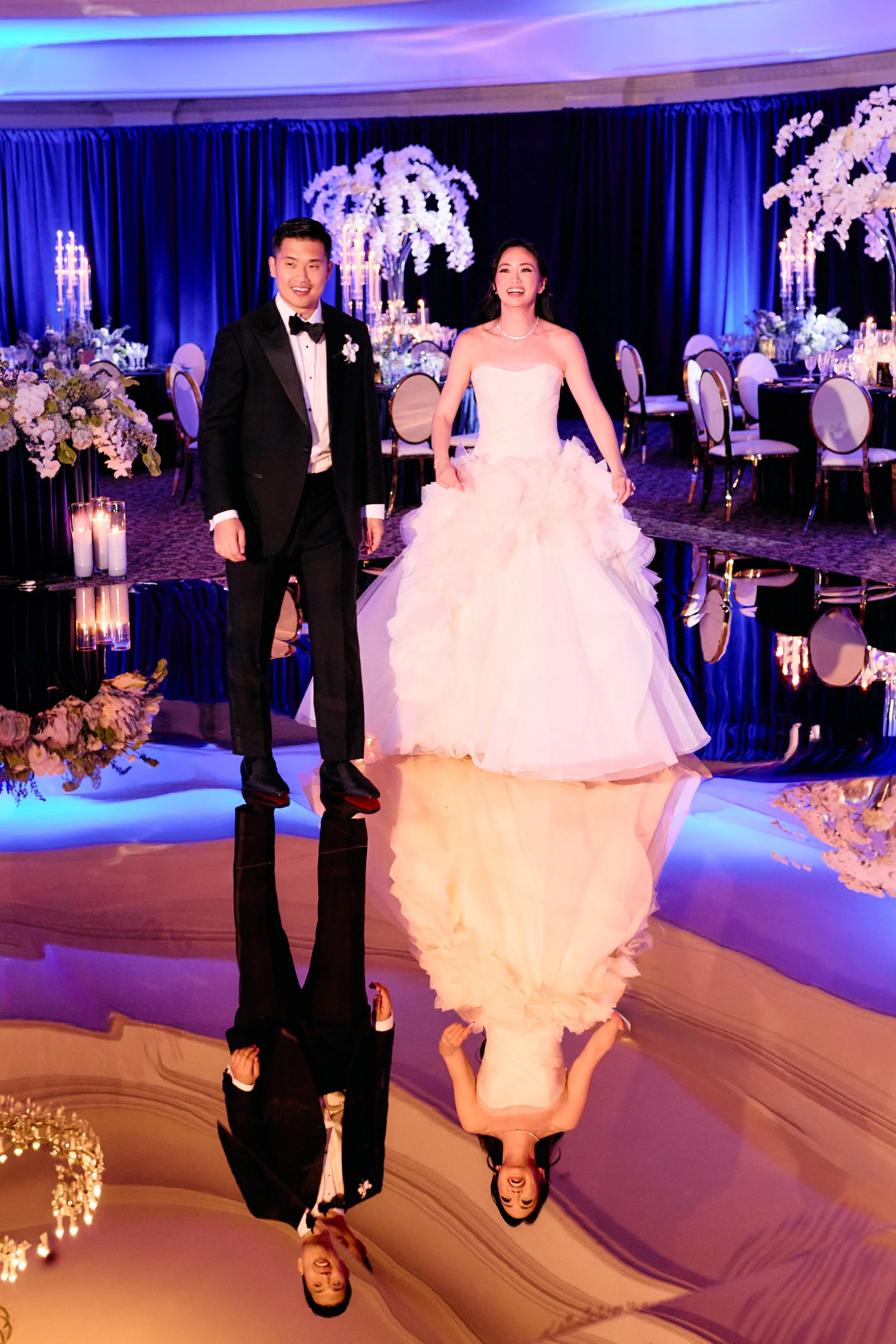 A couple dressed in formal wedding attire standing in a decorated banquet hall with floral arrangements and candles, with a mirror creating a reflection of the scene.