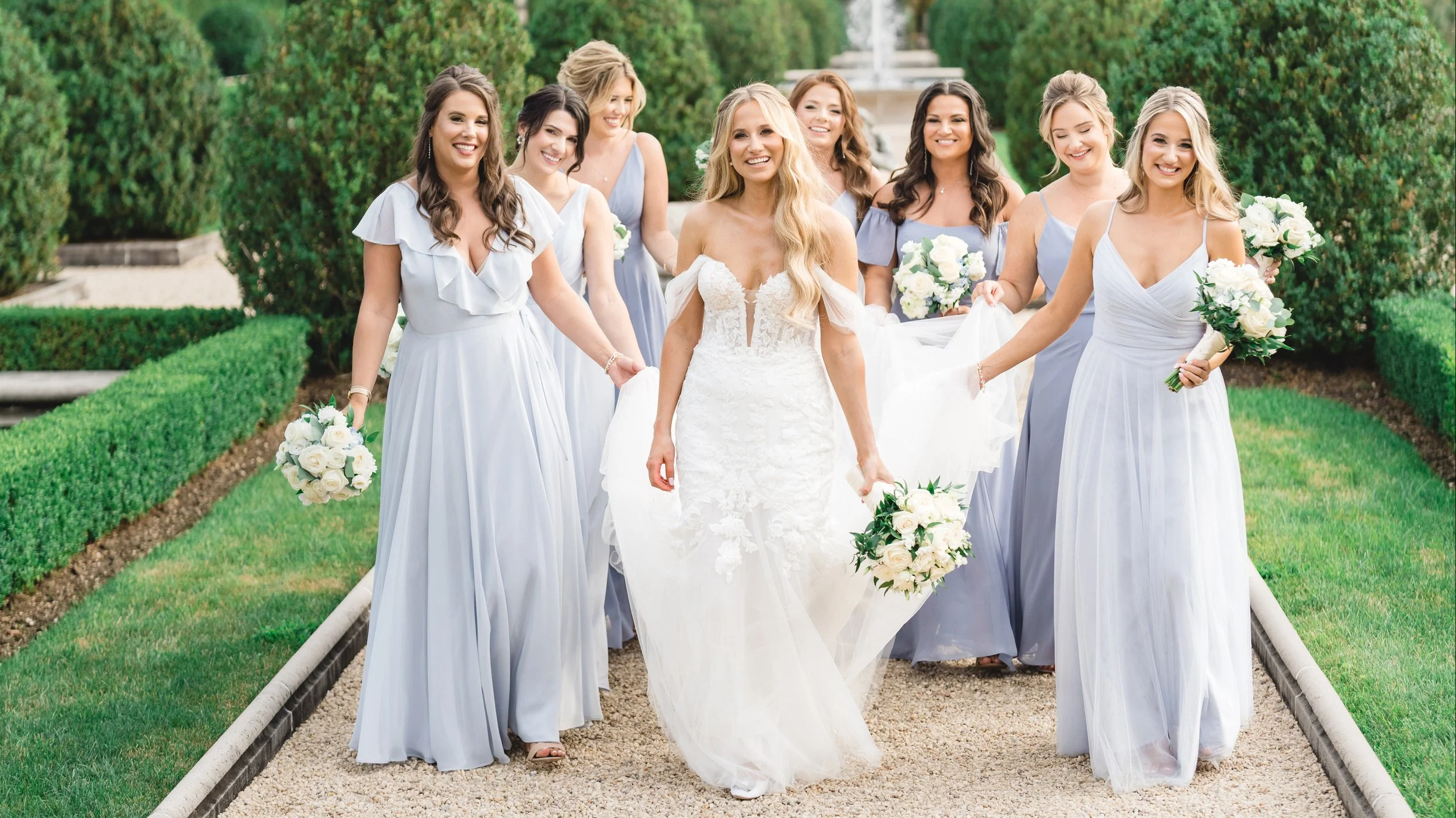 A bride in a white lace wedding dress walking with her bridesmaids holding bouquets of white roses, outdoors in a garden.