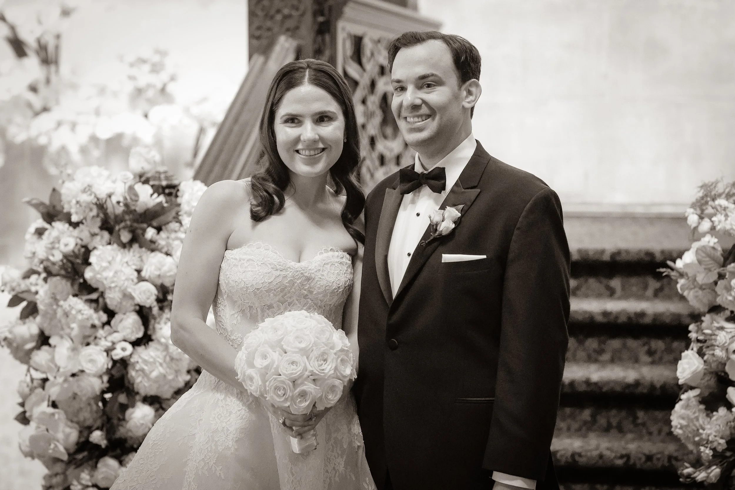 A bride and groom standing together at a wedding, surrounded by floral decorations. The bride is holding a bouquet of roses and wearing a lace wedding dress. The groom is dressed in a tuxedo with a bow tie and boutonniere.