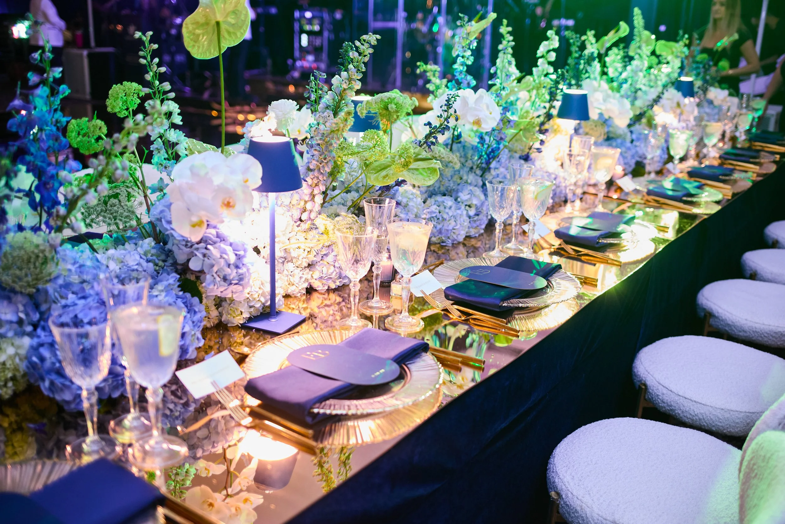 A banquet table decorated with a large arrangement of white and pastel-colored flowers, illuminated by blue lamps and soft lighting, set for a special event or celebration.