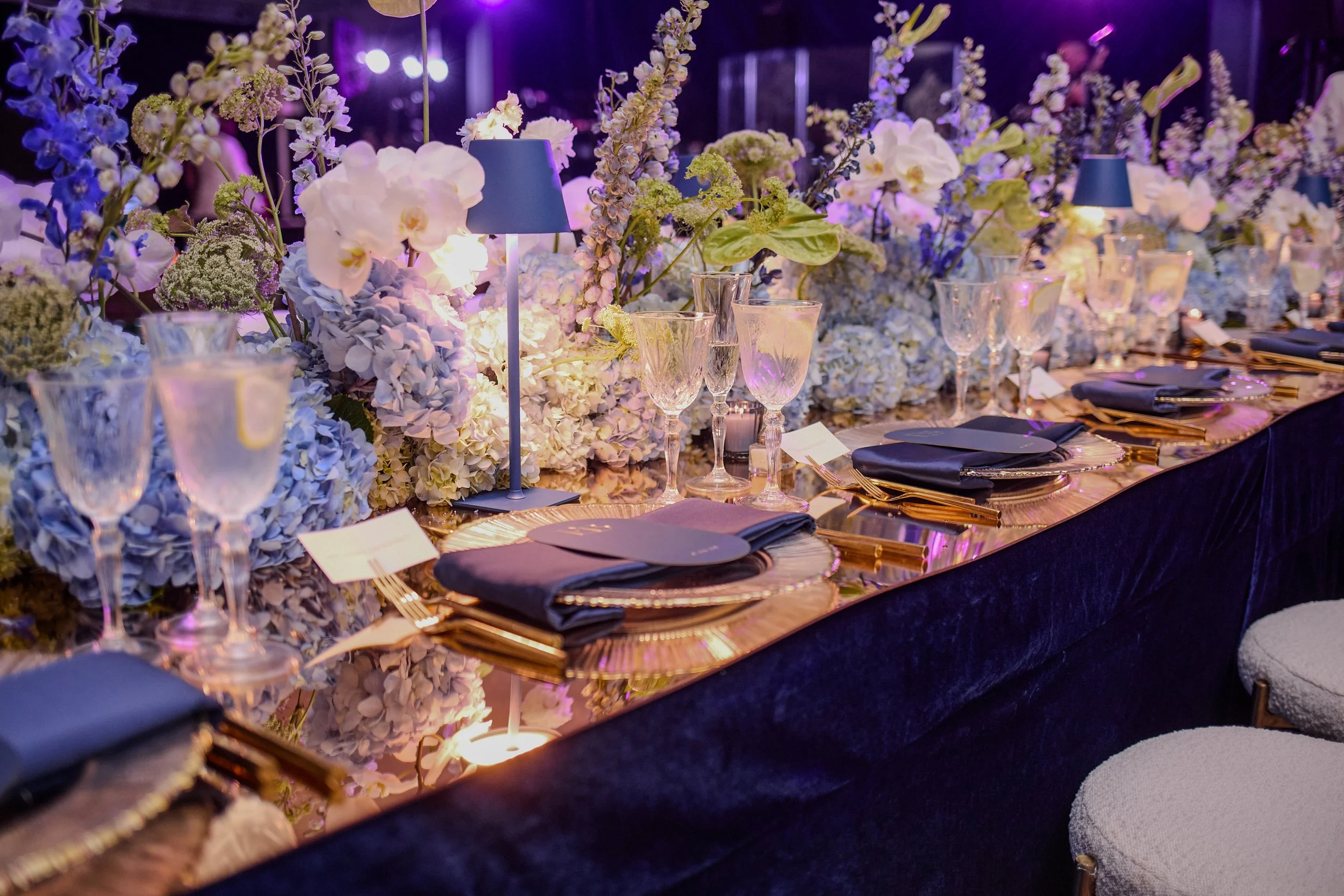 The reception unfolded in Oheka Castle’s Terrace Room, where king’s tables were designed in rich layers of deep blue, gold, and soft candlelight. Lush arrangements of hydrangea and delicate florals spilled across mirrored surfaces, creating a sense o