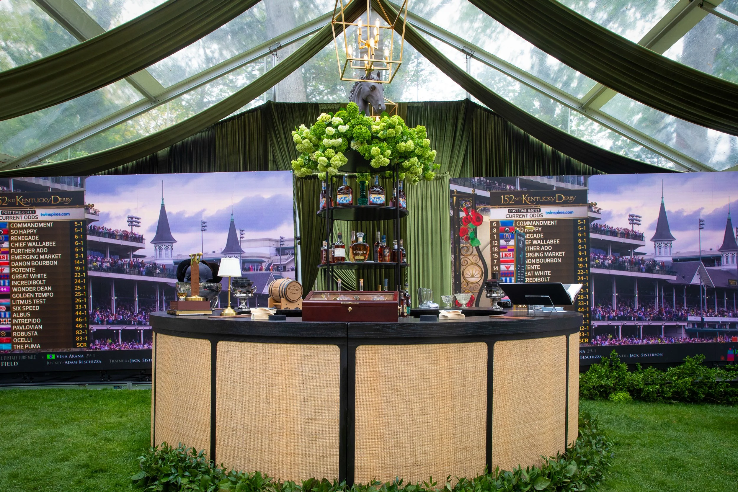 A bespoke bourbon bar featuring live Kentucky Derby race coverage, custom shelving, and layered greenery beneath the tented reception space.