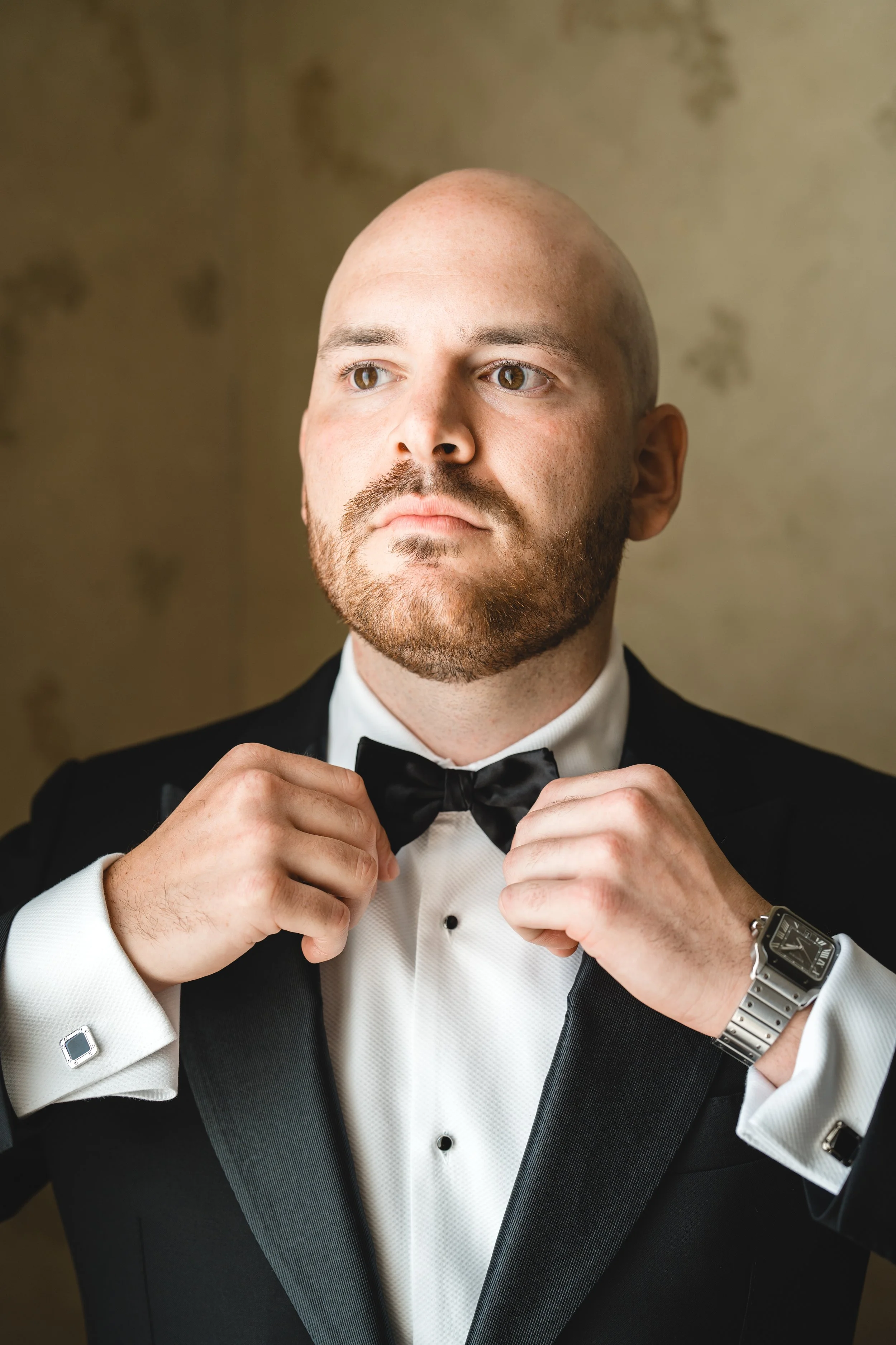 Groom adjusting black bow tie in tuxedo with soft window light portrait